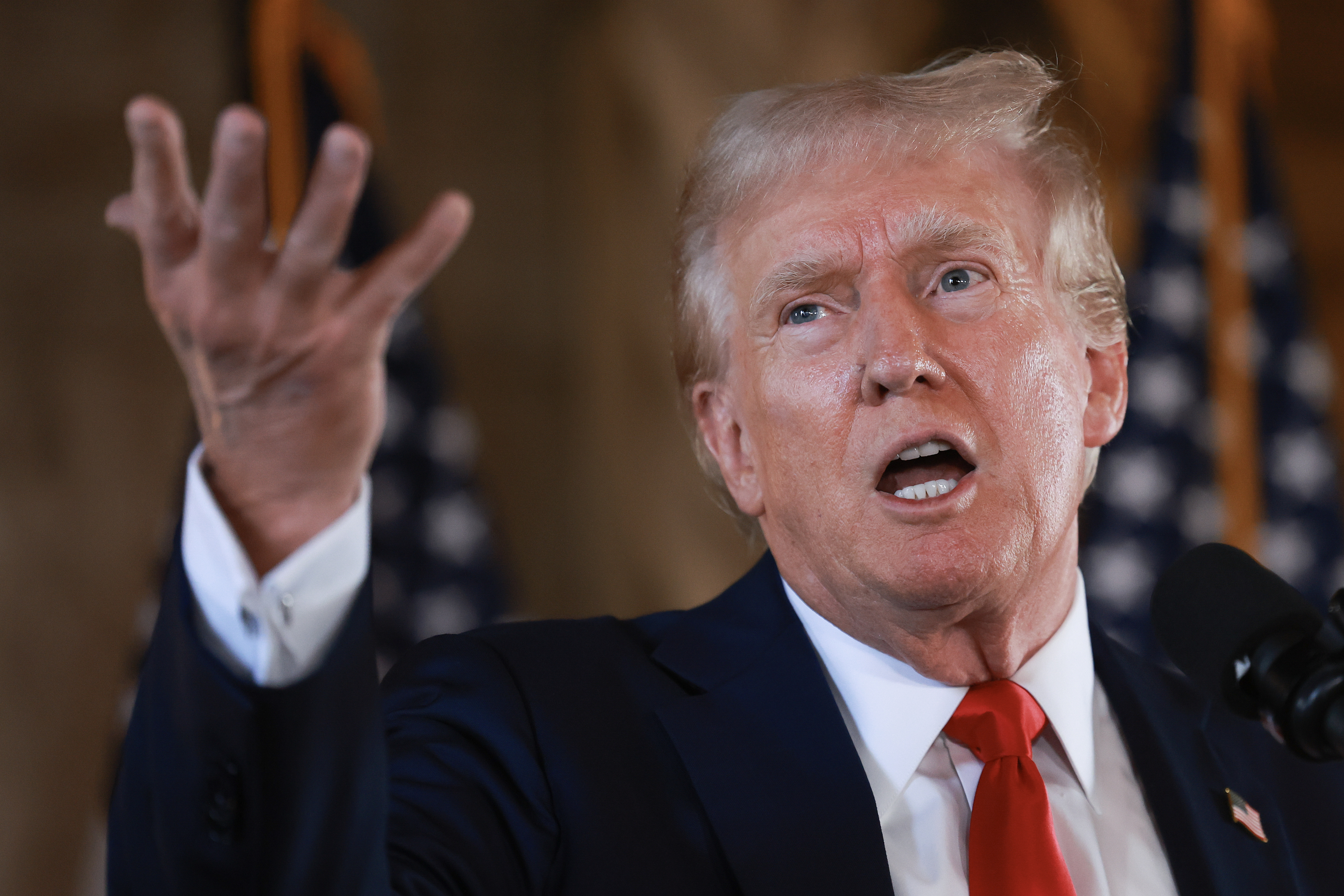 A man in a suit and red tie, gesturing with his left hand while speaking at a podium, with American flags in the background