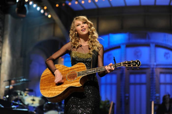 Taylor Swift performing on stage with a guitar, wearing a sparkly black dress. The background has a lit stage and band members