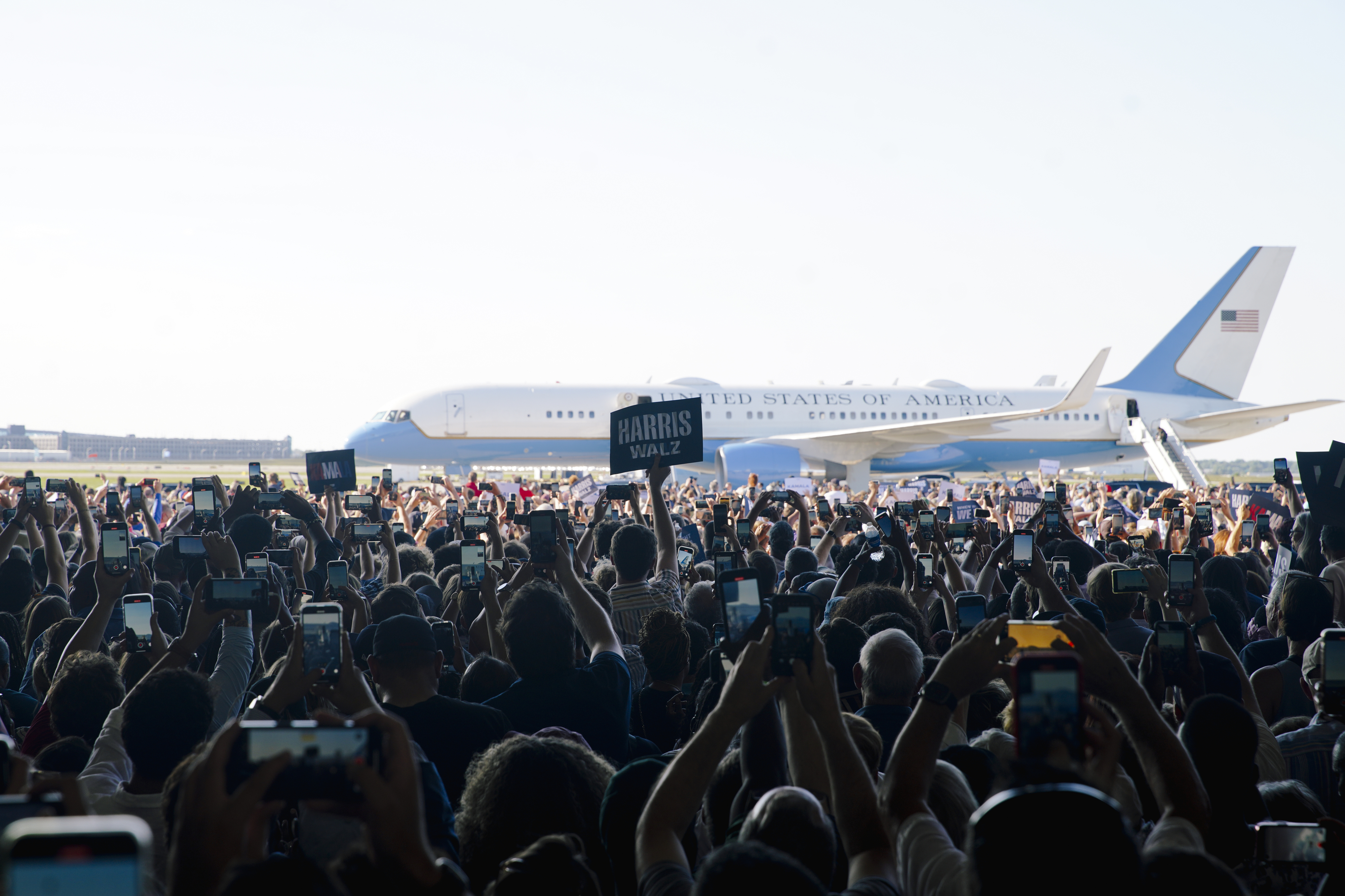 A large crowd holds up phones to take photos of Air Force One at an airfield event. Signs reading "Harris" and "Biden" are visible among the crowd