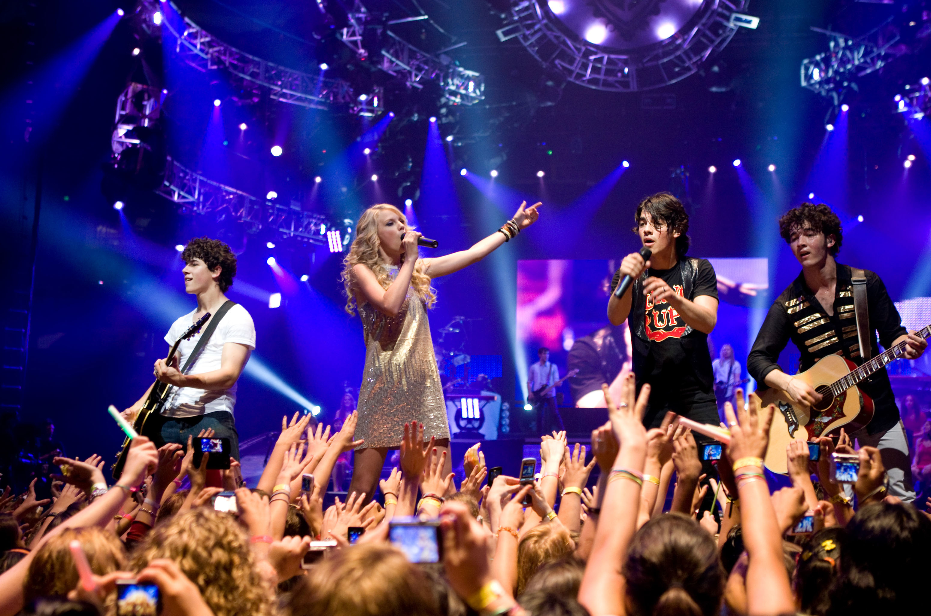 Nick Jonas, Taylor Swift, Joe Jonas, and Kevin Jonas perform on stage in front of an energetic crowd, with Taylor pointing and singing into the microphone