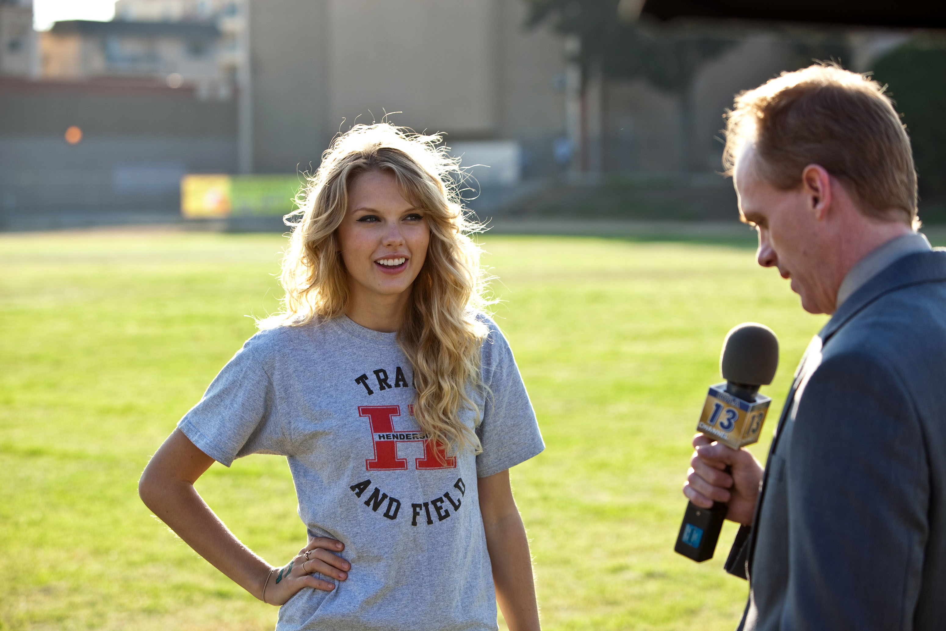 Taylor Swift, wearing a &quot;Track and Field&quot; t-shirt, is being interviewed by a man in a suit holding a microphone with a &quot;13&quot; emblem