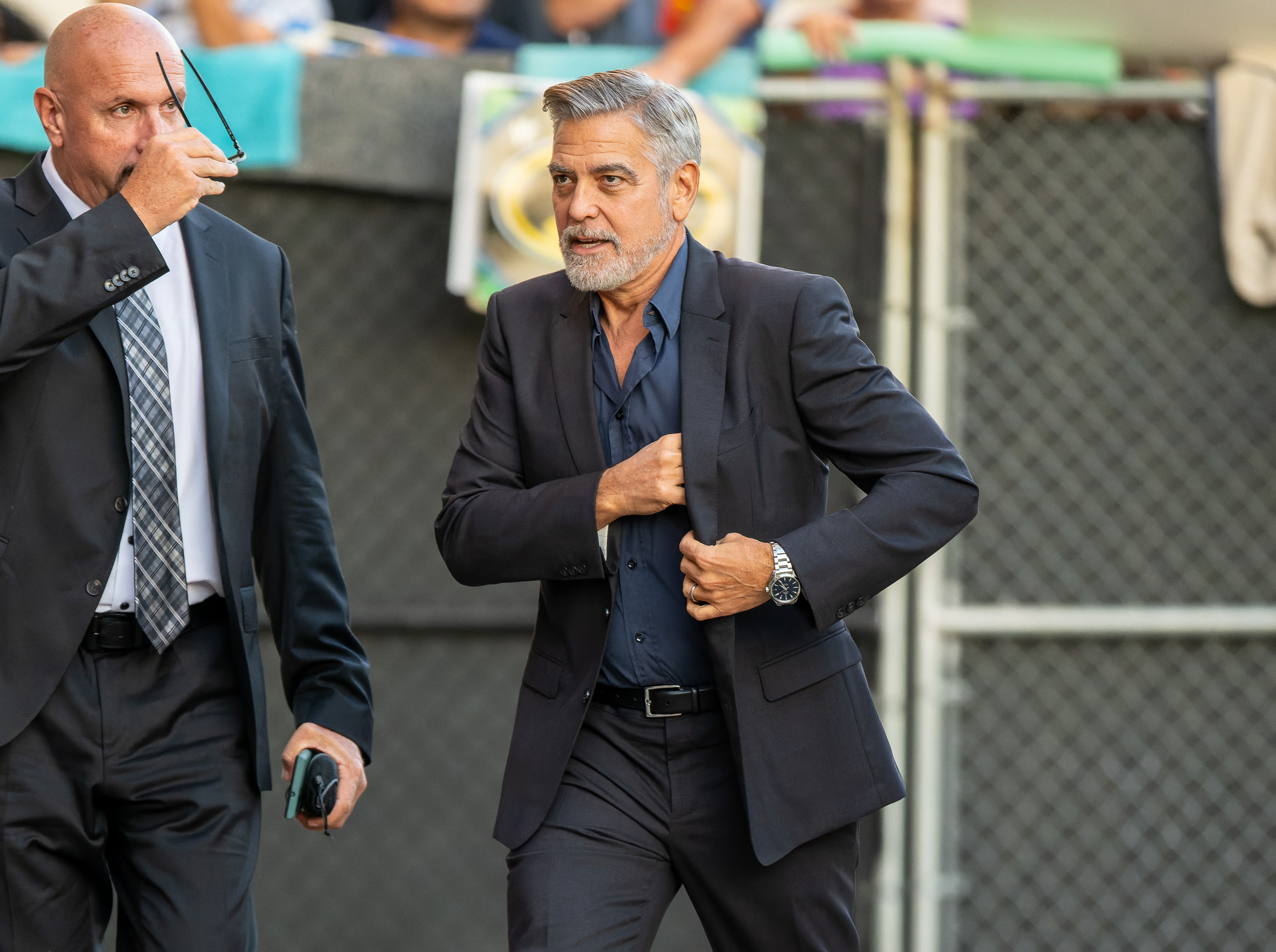 George Clooney in a dark suit adjusts his jacket while walking with a suited man