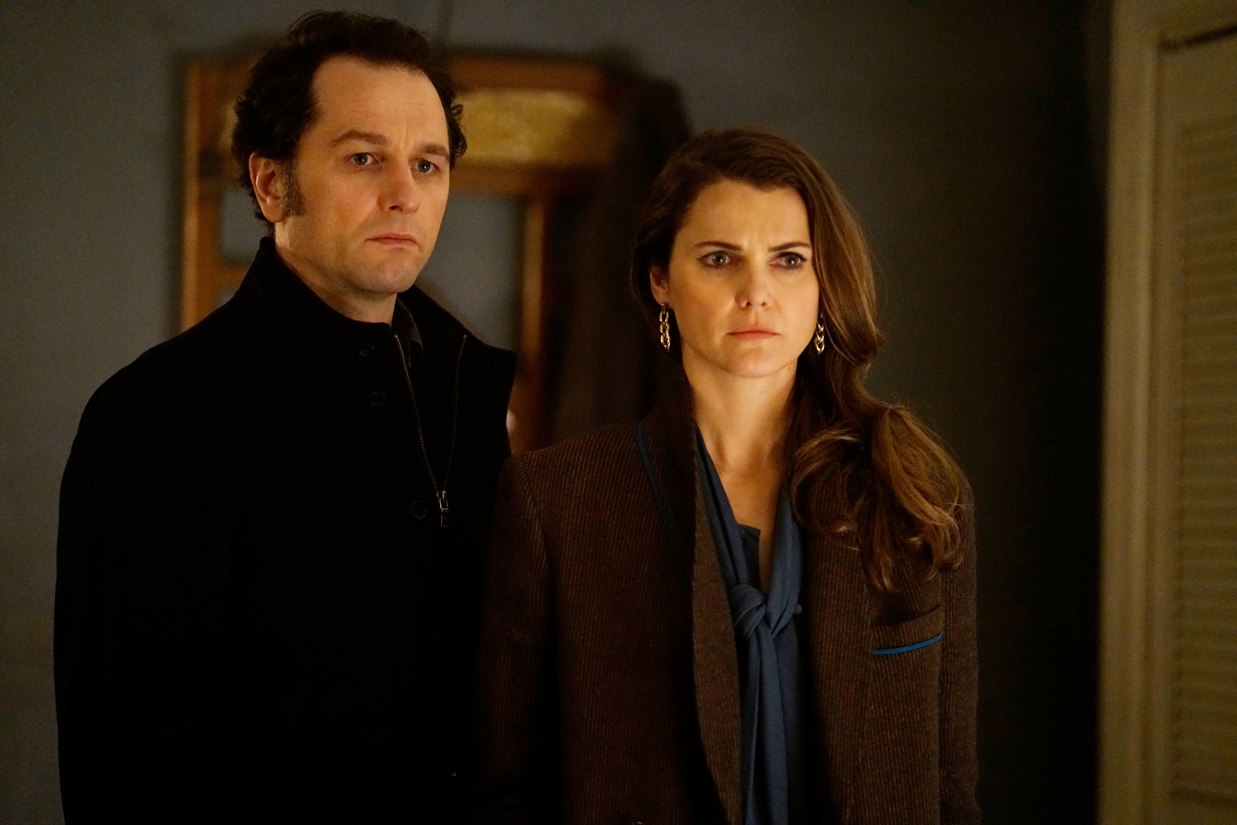 Matthew Rhys and Keri Russell stand side by side in formal attire, looking serious, in a scene from a TV show
