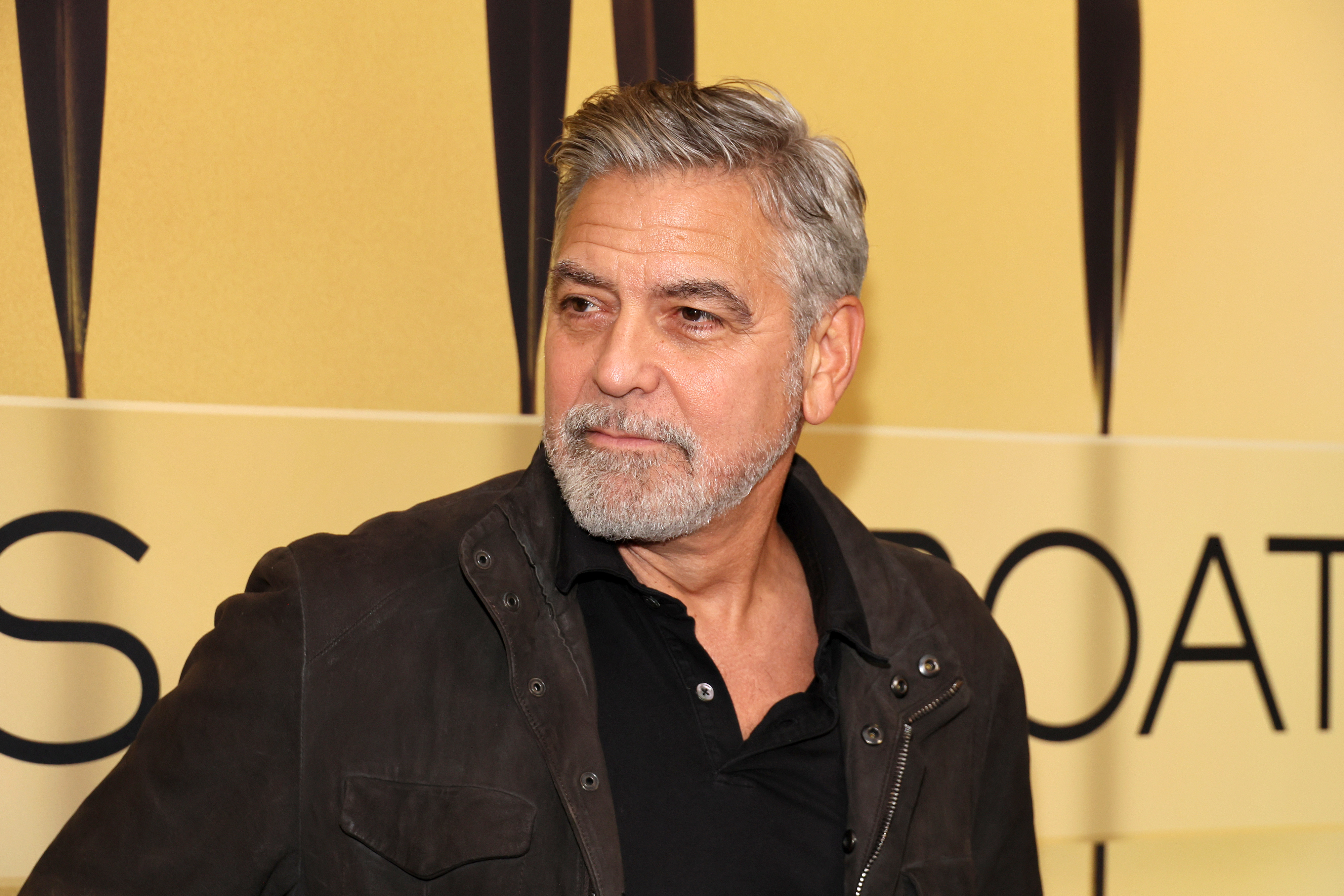George Clooney attends an event wearing a dark jacket over a black shirt, with a serious expression