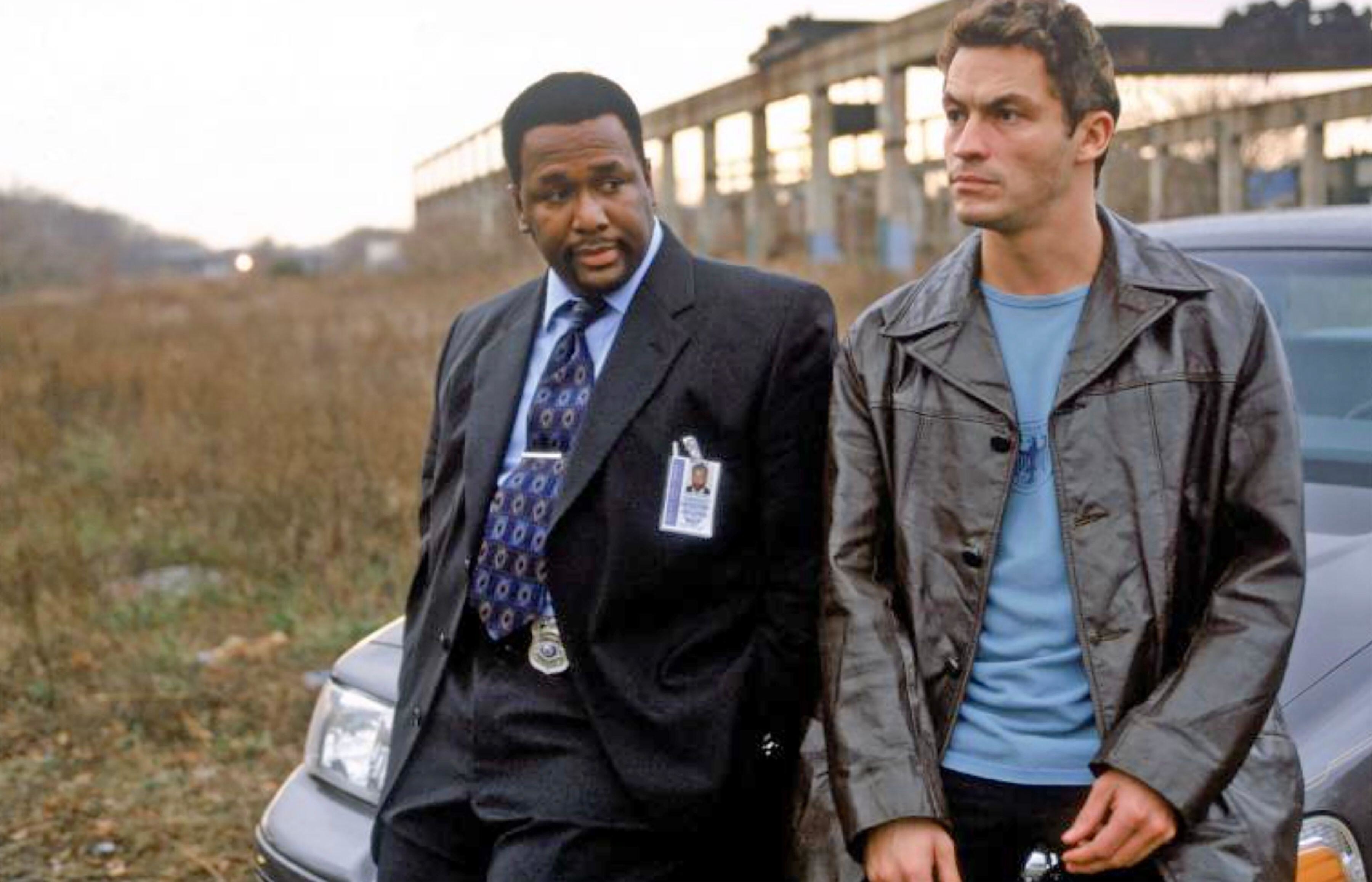 Wendell Pierce in a suit and tie, and Dominic West in a casual jacket, standing outdoors next to a car, possibly discussing something