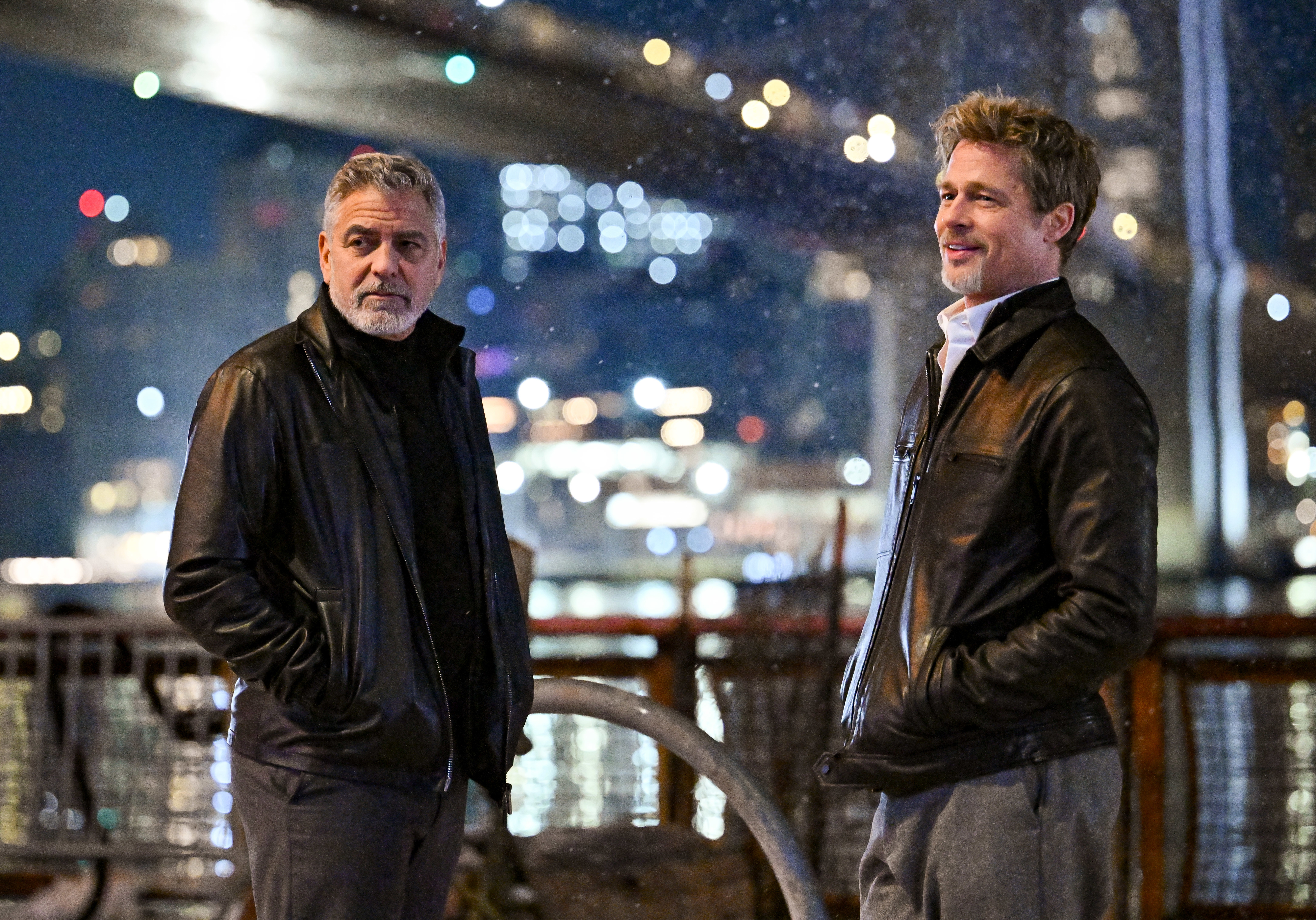 George Clooney and Brad Pitt are standing outdoors at night, both wearing leather jackets. They are smiling and appear to be engaged in a conversation