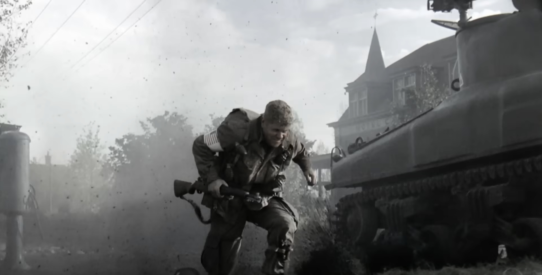 A soldier crouches and runs in front of a tank in a bombed-out WWII village, dust clouds and rubble all around