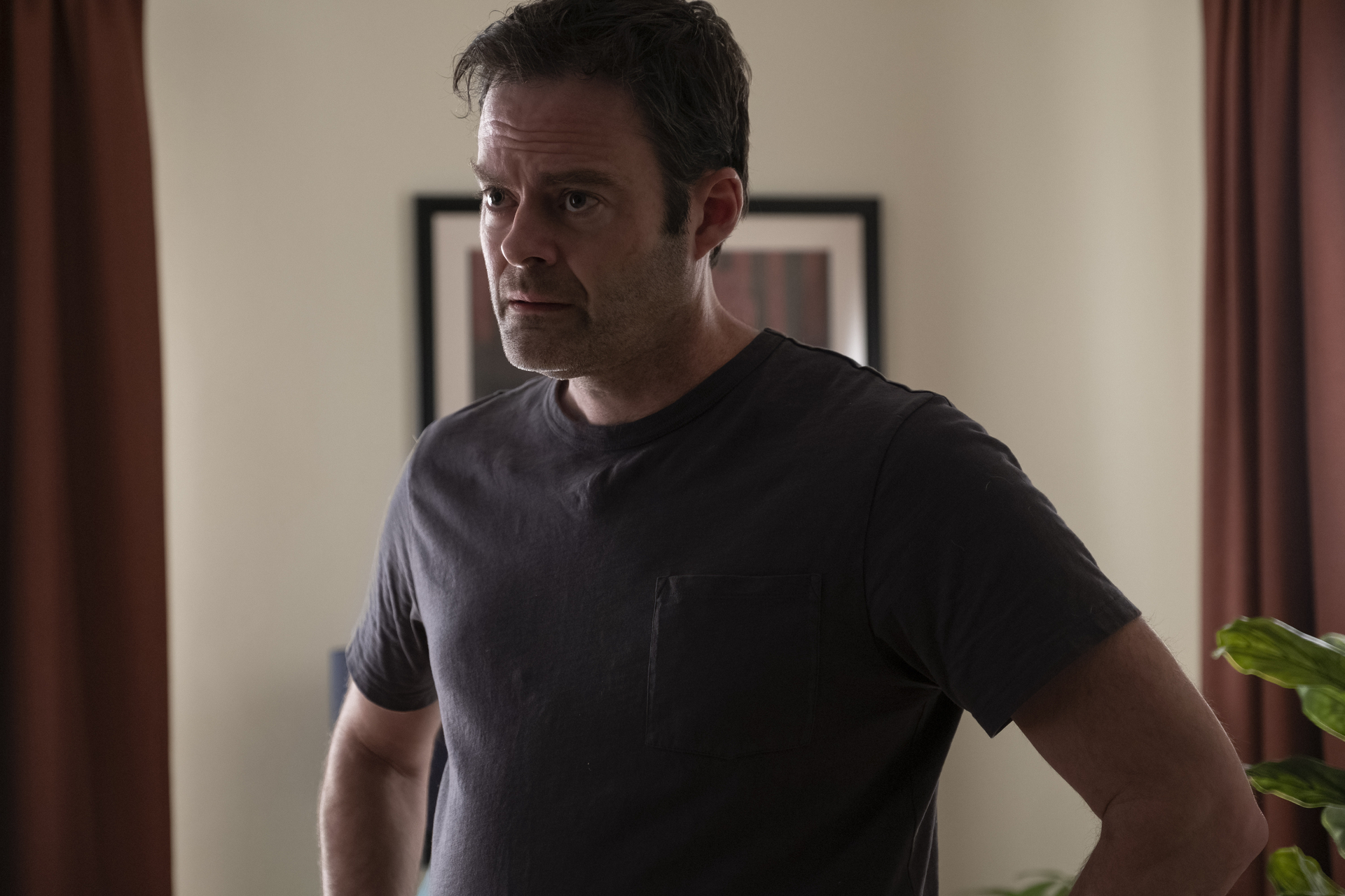Bill Hader in a plain t-shirt stands with hands on hips, looking contemplative in an indoor setting