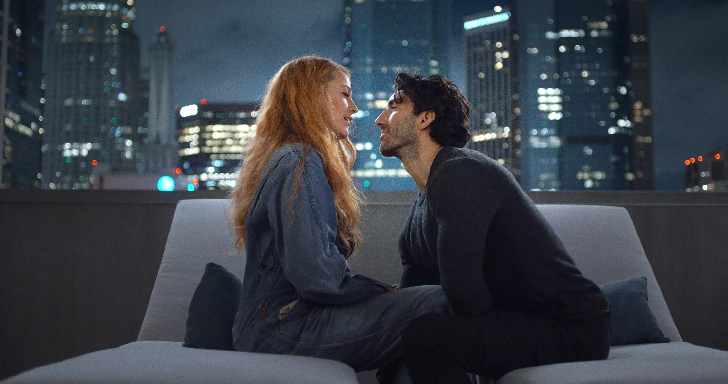 A man and a woman sit on a rooftop couch at night, face to face and smiling, with a city skyline in the background. Names unknown