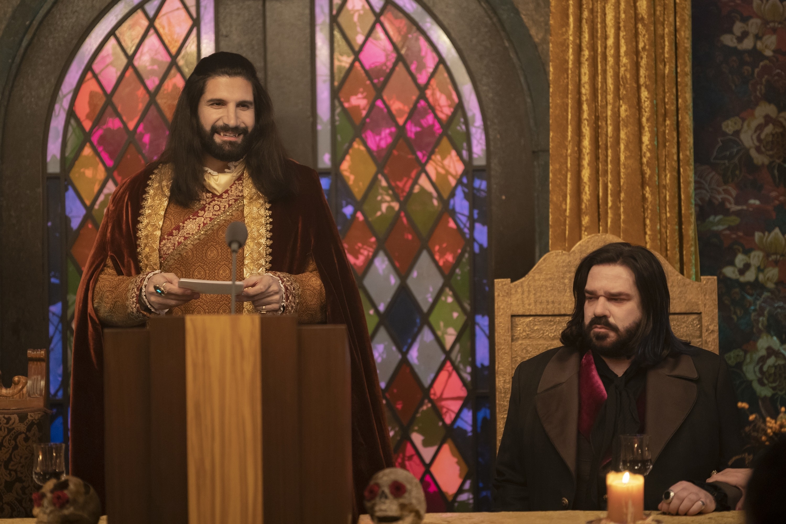 Kayvan Novak speaking at a podium while Matt Berry sits beside him at a table in a scene from the TV show &quot;What We Do in the Shadows&quot;