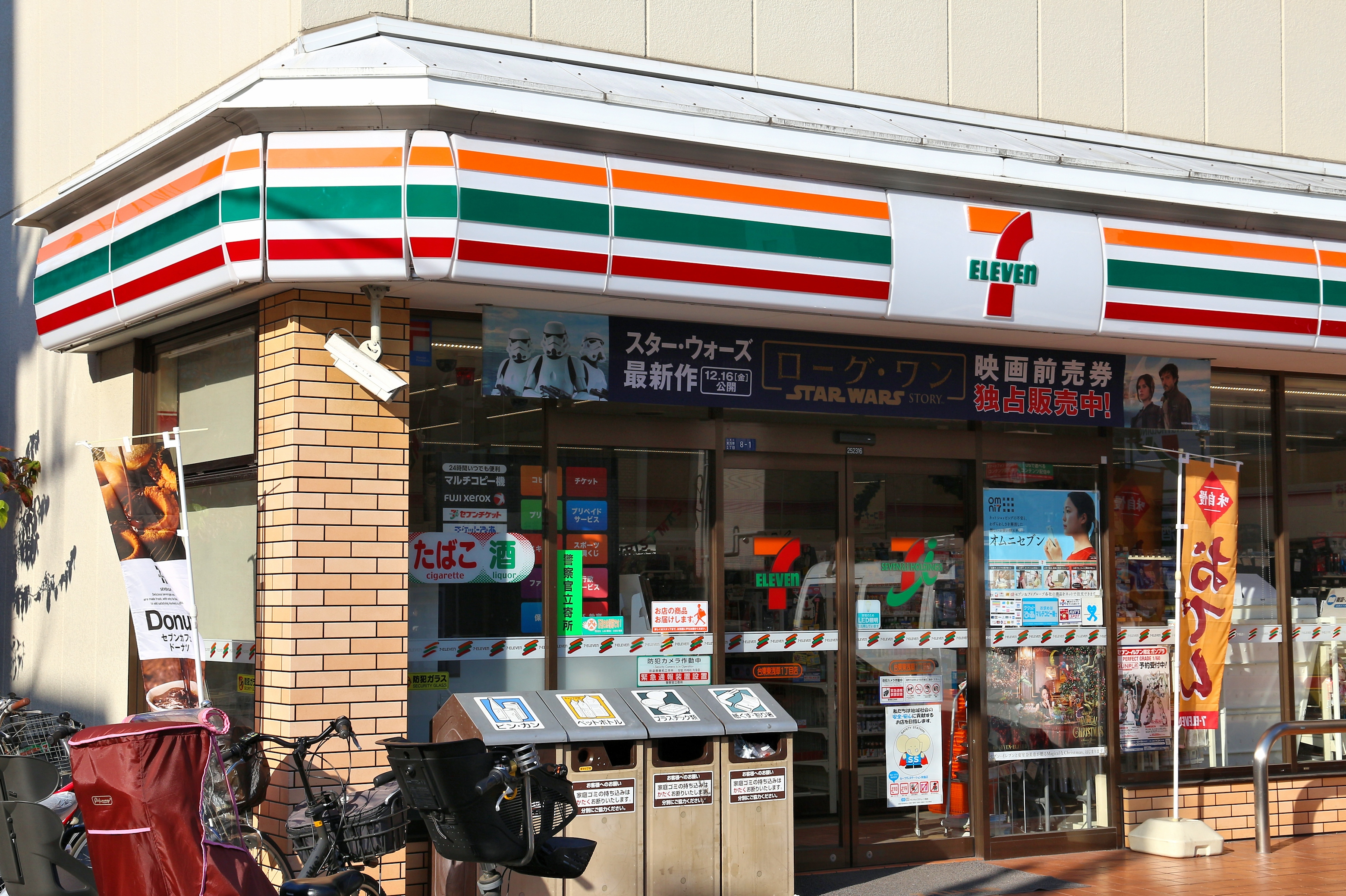 A 7-Eleven store entrance in Japan. Bicycles are parked outside, and various promotional posters are visible through the windows
