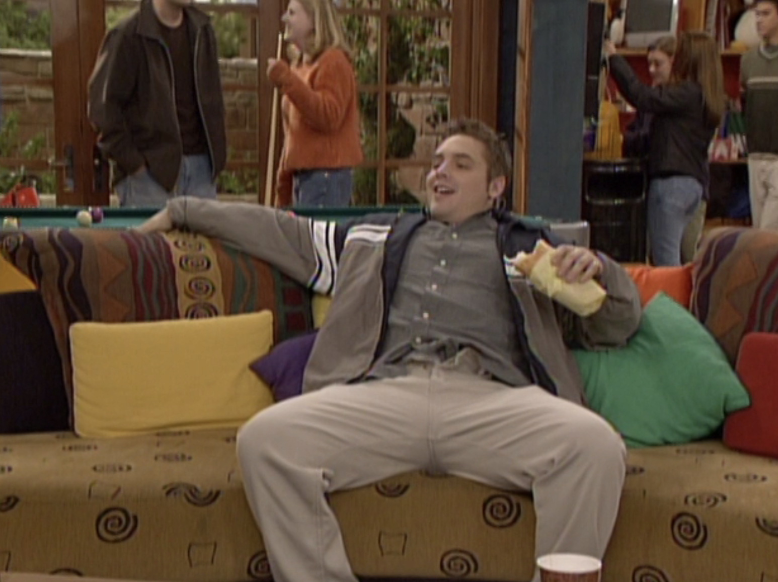 Will Friedle seated on a couch with colorful pillows, holding a burrito and smiling. Other people socialize in the background at what appears to be a casual gathering