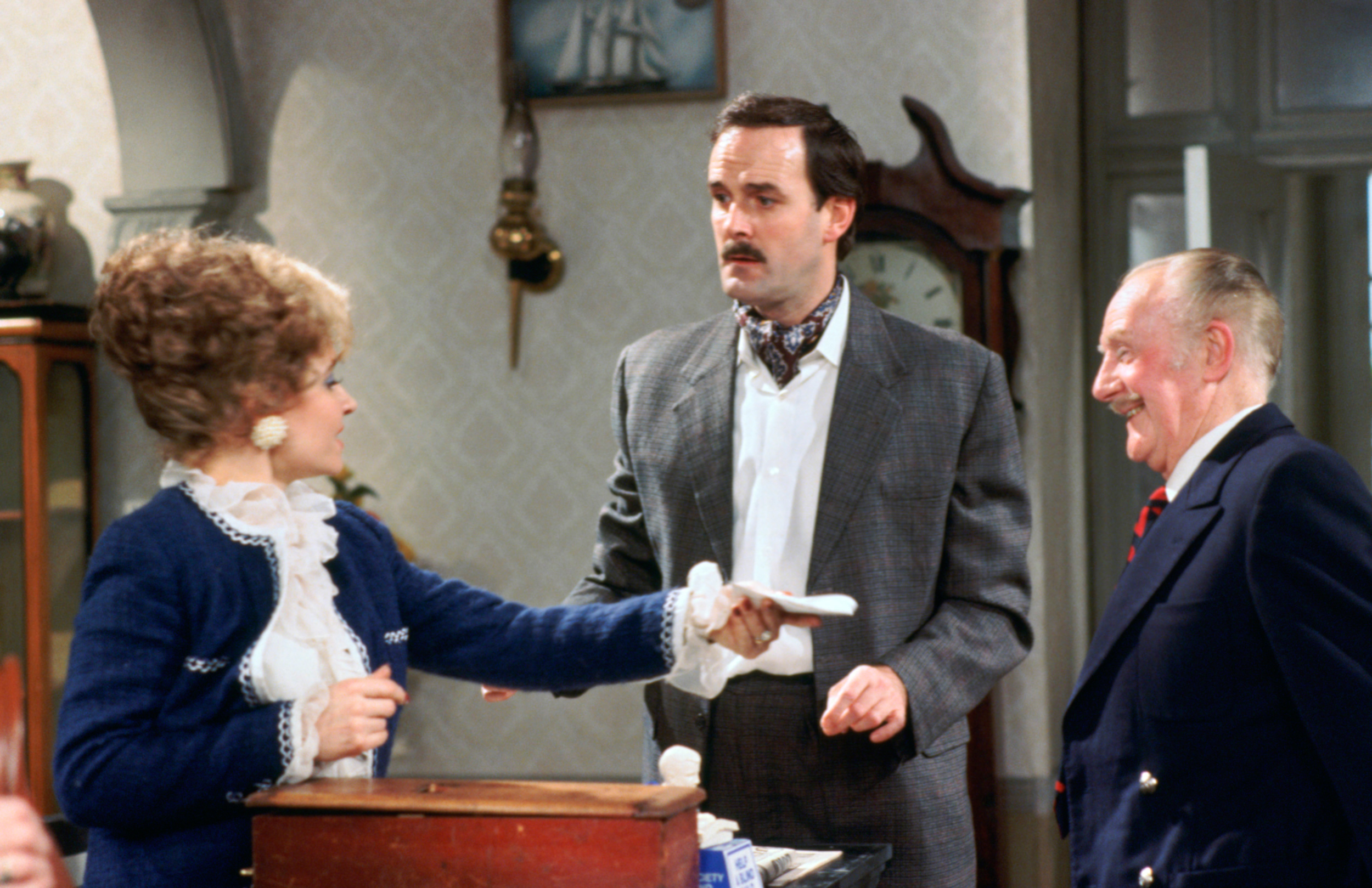 Connie Booth, John Cleese, and Andrew Sachs in a scene from a classic TV show. Booth holds out a paper as Cleese and Sachs look at her, appearing in period attire