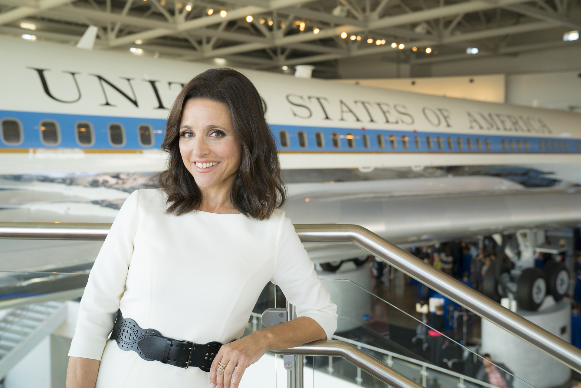 Julia Louis-Dreyfus smiles while leaning on a railing with a backdrop of a &quot;United States of America&quot; airplane in an indoor exhibition. She wears a white dress with a black belt