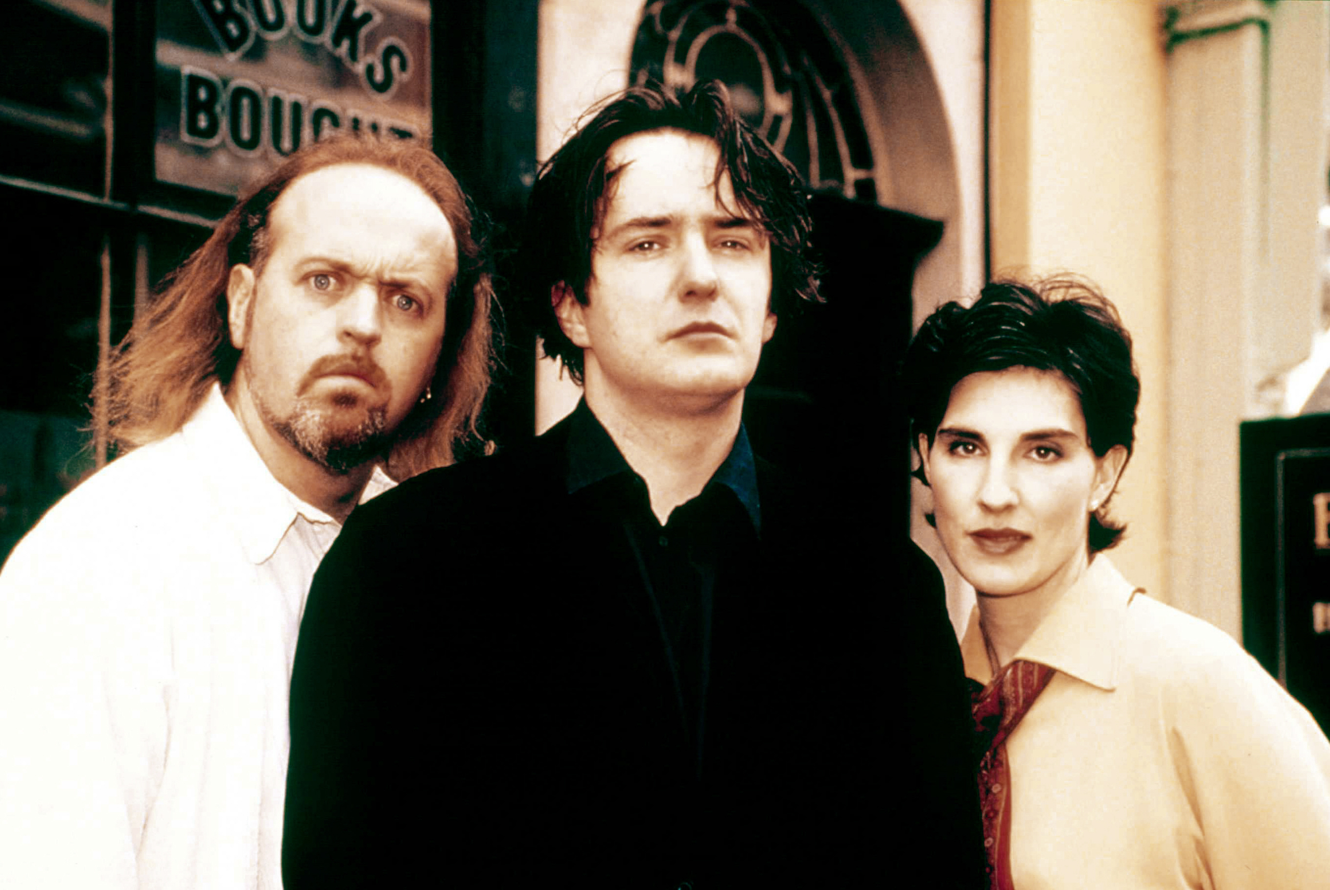 Bill Bailey, Dylan Moran, and Tamsin Greig stand outside a bookshop. Bailey has a confused expression, Moran looks serious, and Greig is smiling slightly