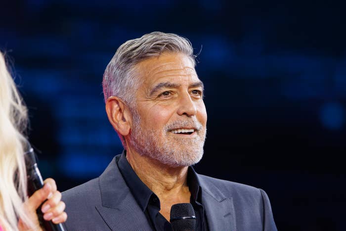 George Clooney, in a tailored suit, smiles and holds a microphone on stage