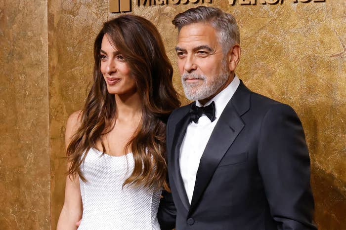 George Clooney in a black tuxedo and Amal Clooney in a white gown pose on a red carpet at a formal event