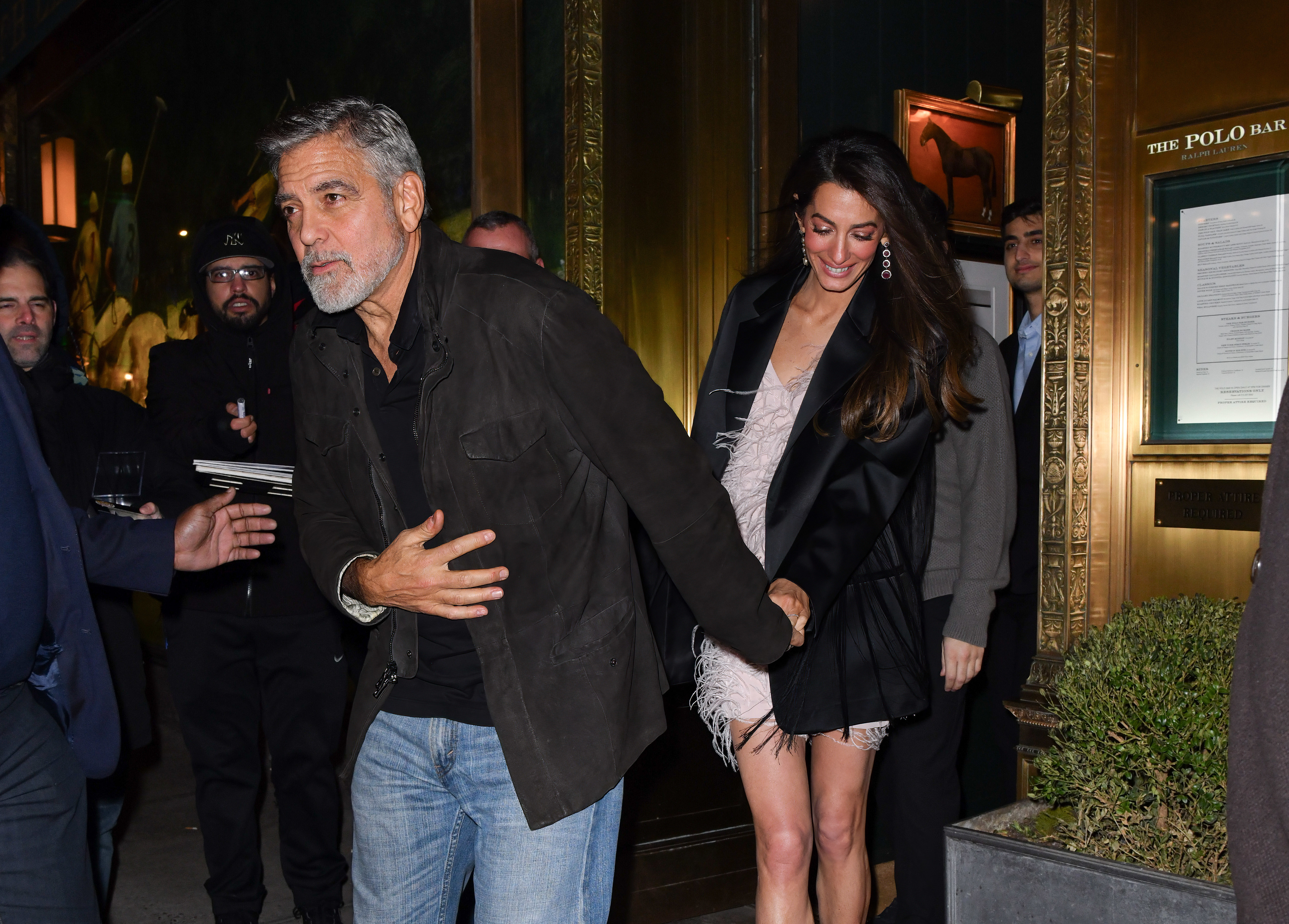 George Clooney and Amal Clooney walk hand in hand outside a building. George is in casual attire, and Amal wears a fashionable short dress with a long coat