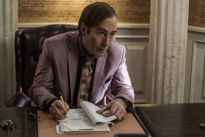 Bob Odenkirk as Saul Goodman in an office, wearing a suit and tie, writing on a notepad. Behind him is a wall with text