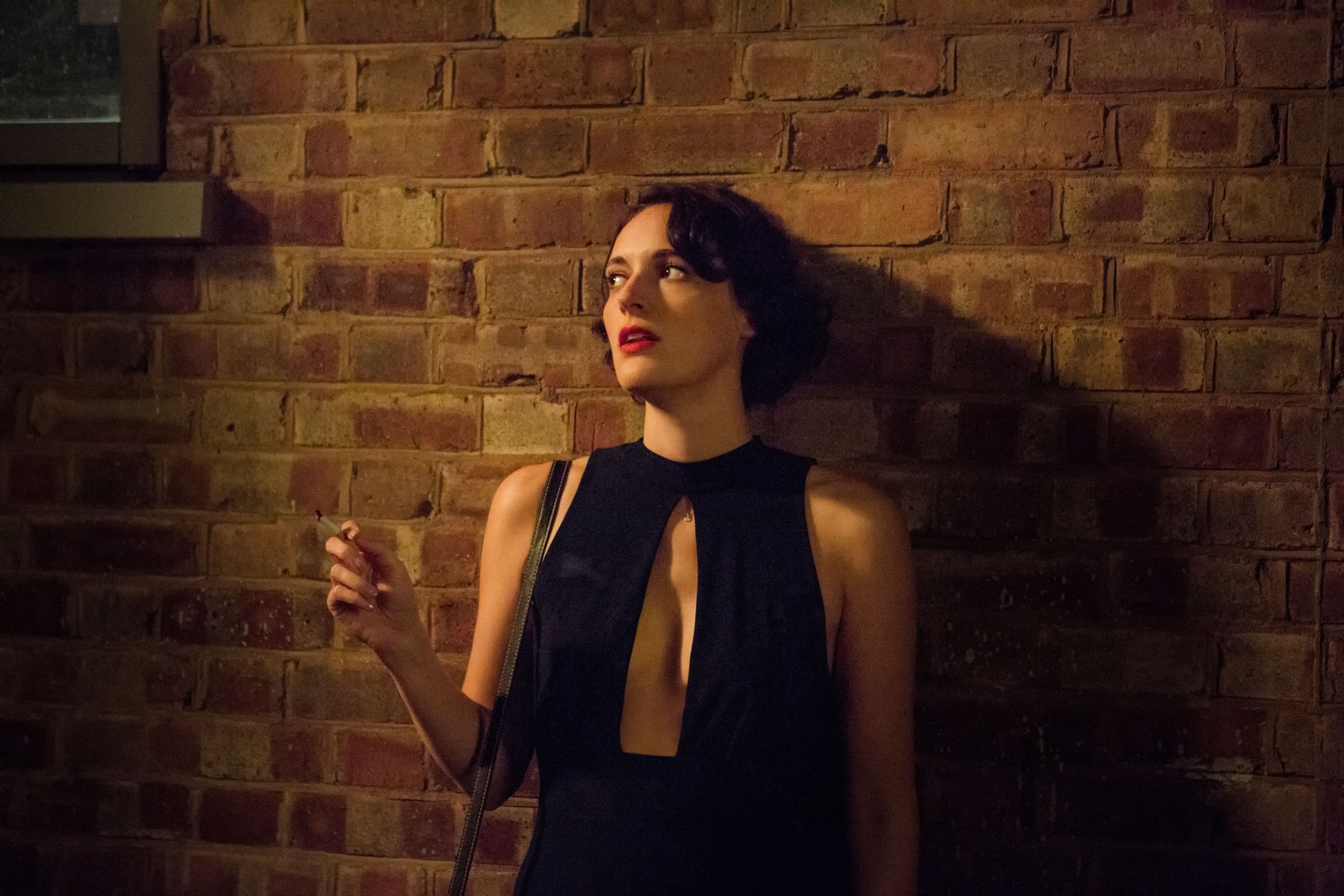 Phoebe Waller-Bridge in a black sleeveless dress with a front cutout, leaning against a brick wall and holding a cigarette