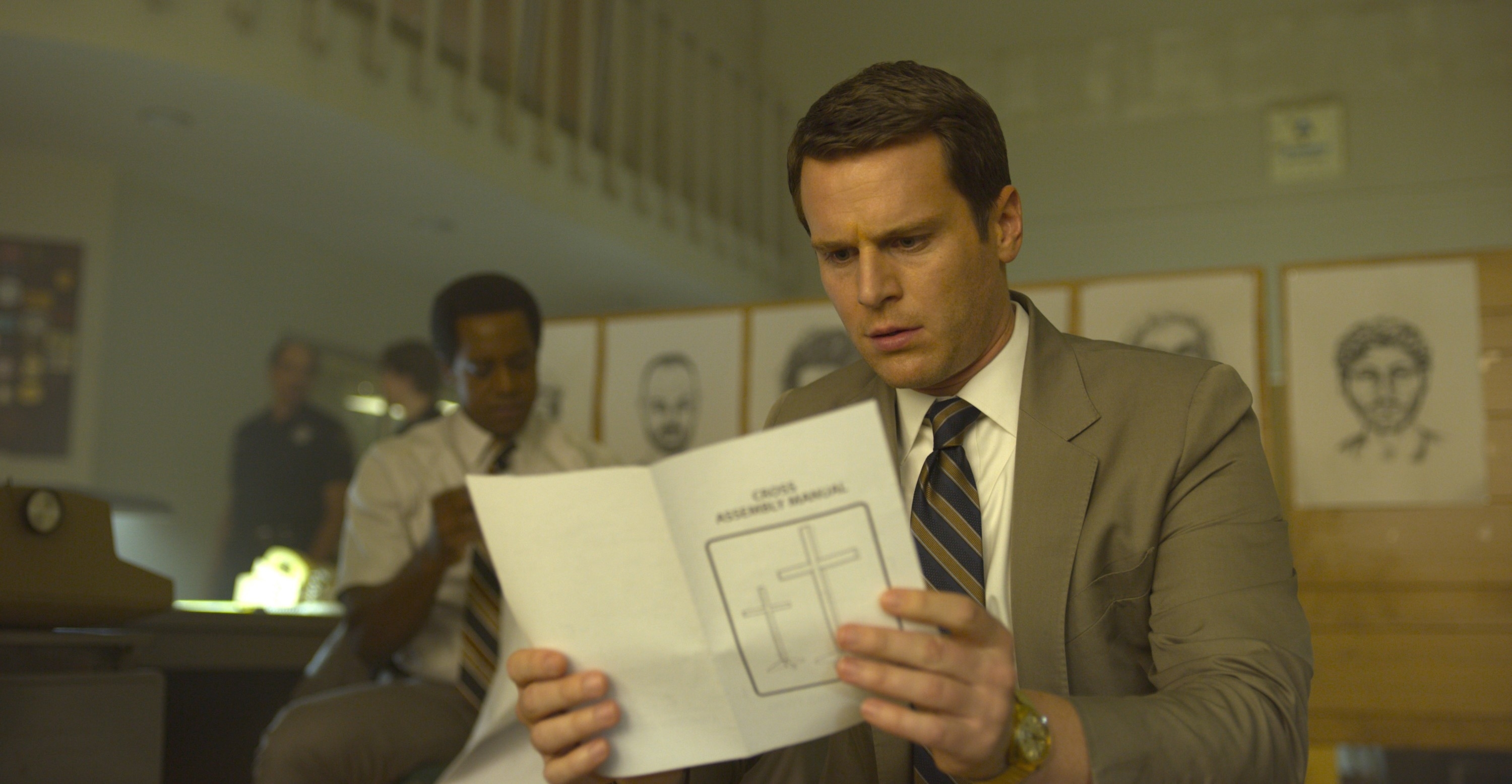 Jonathan Groff and Holt McCallany review a document in an office setting with crime sketches on the wall