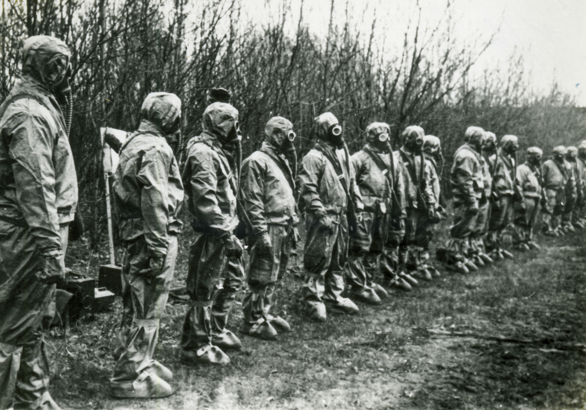 A group of people stand in a line wearing full-body hazmat suits and gas masks in a forested area