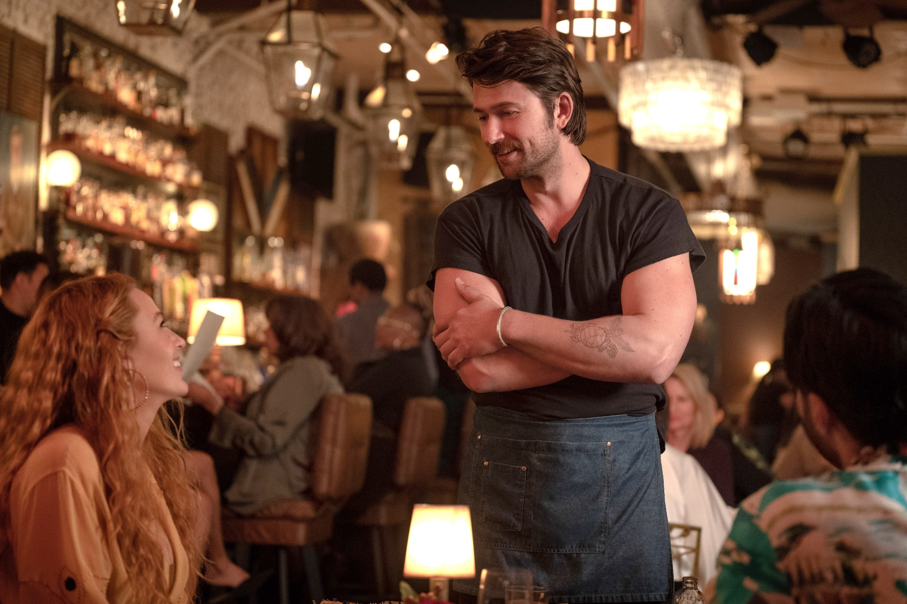 Blake lively (left) talks to Brandon Sklenar (center) in a busy bar. He is wearing a dark t-shirt and an apron, standing with his arms crossed