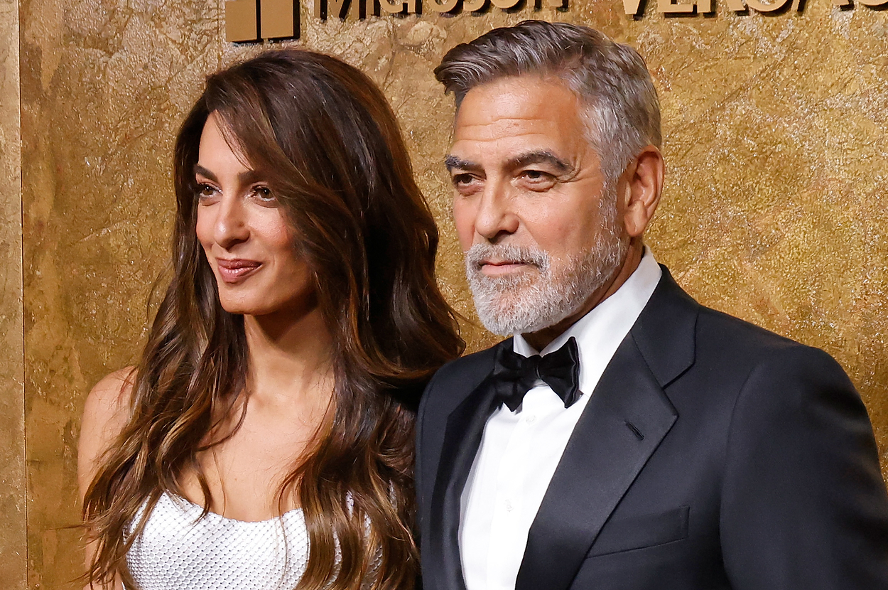 George Clooney in a black tuxedo and Amal Clooney in a white gown pose on a red carpet at a formal event