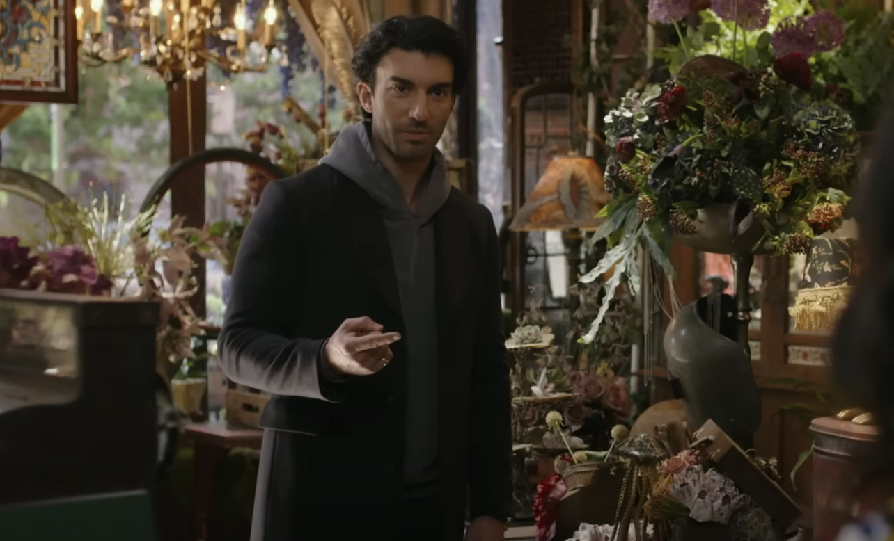 Justin Baldoni stands in a flower shop, wearing a dark coat over a hoodie, in a scene from a TV show or movie