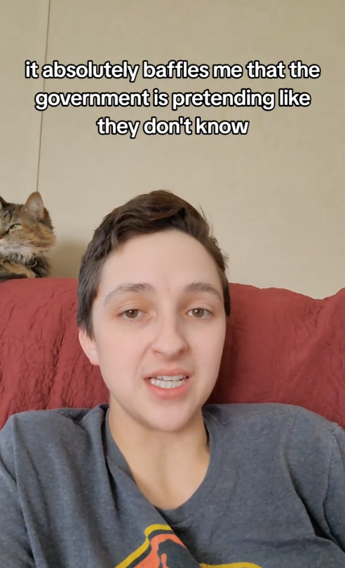Person in a casual shirt speaking on a red couch with a cat behind them; text reads, "it absolutely baffles me that the government is pretending like they don't know."