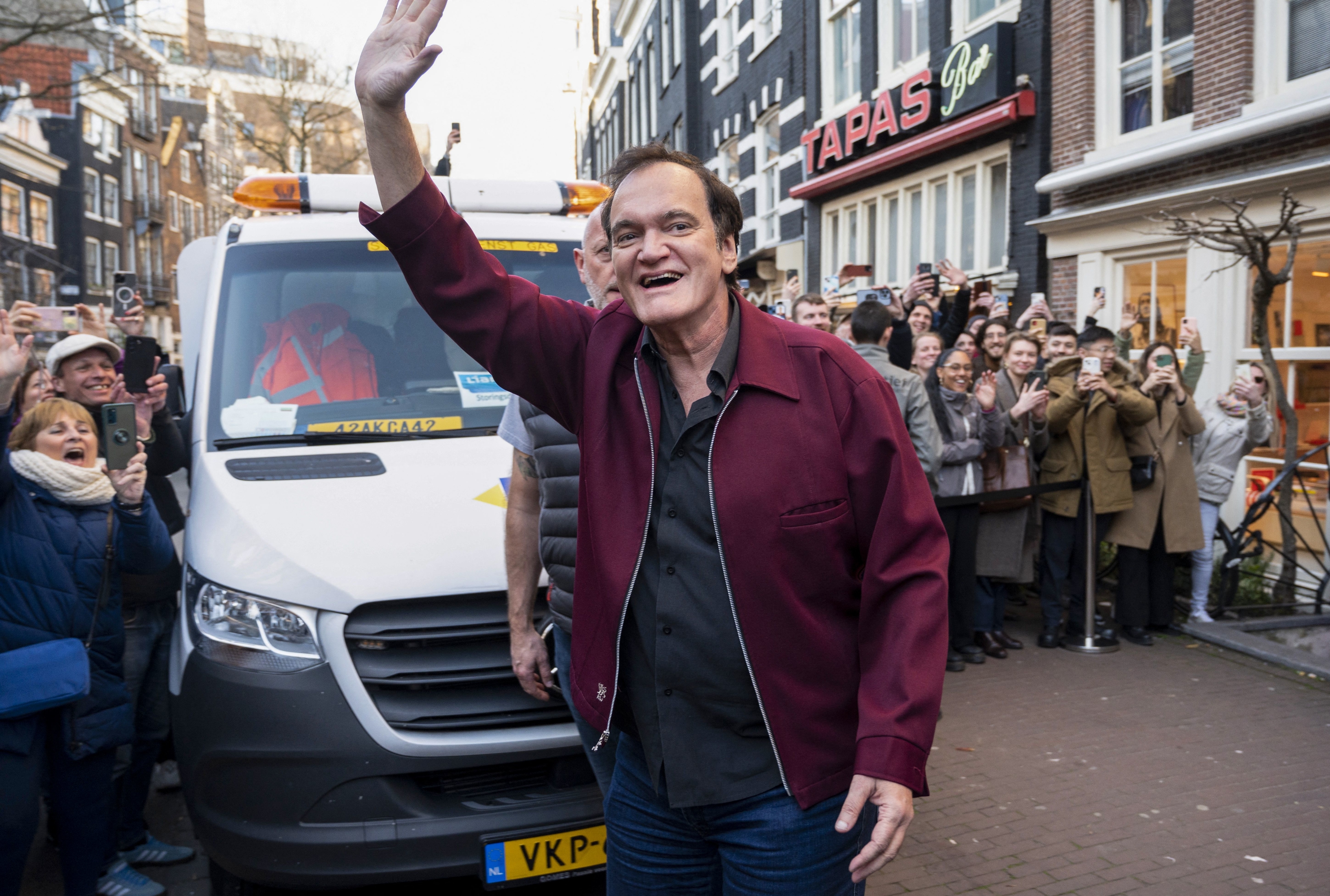 Quentin Tarantino waves at fans while standing in front of a van. Fans around take photos and cheer