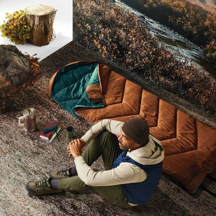 The sleeping bag is laid flat next to the model in an outdoor setting, near a stream