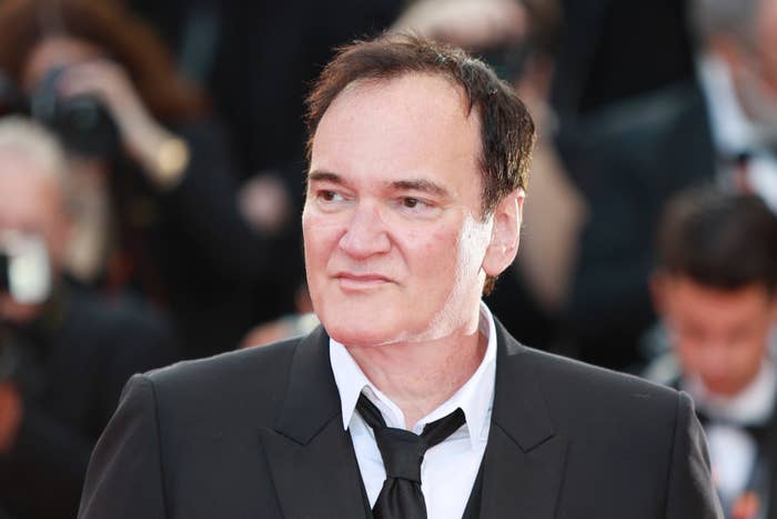 Quentin Tarantino on the red carpet, wearing a suit and tie, looking slightly to the side. Background shows a blurred crowd