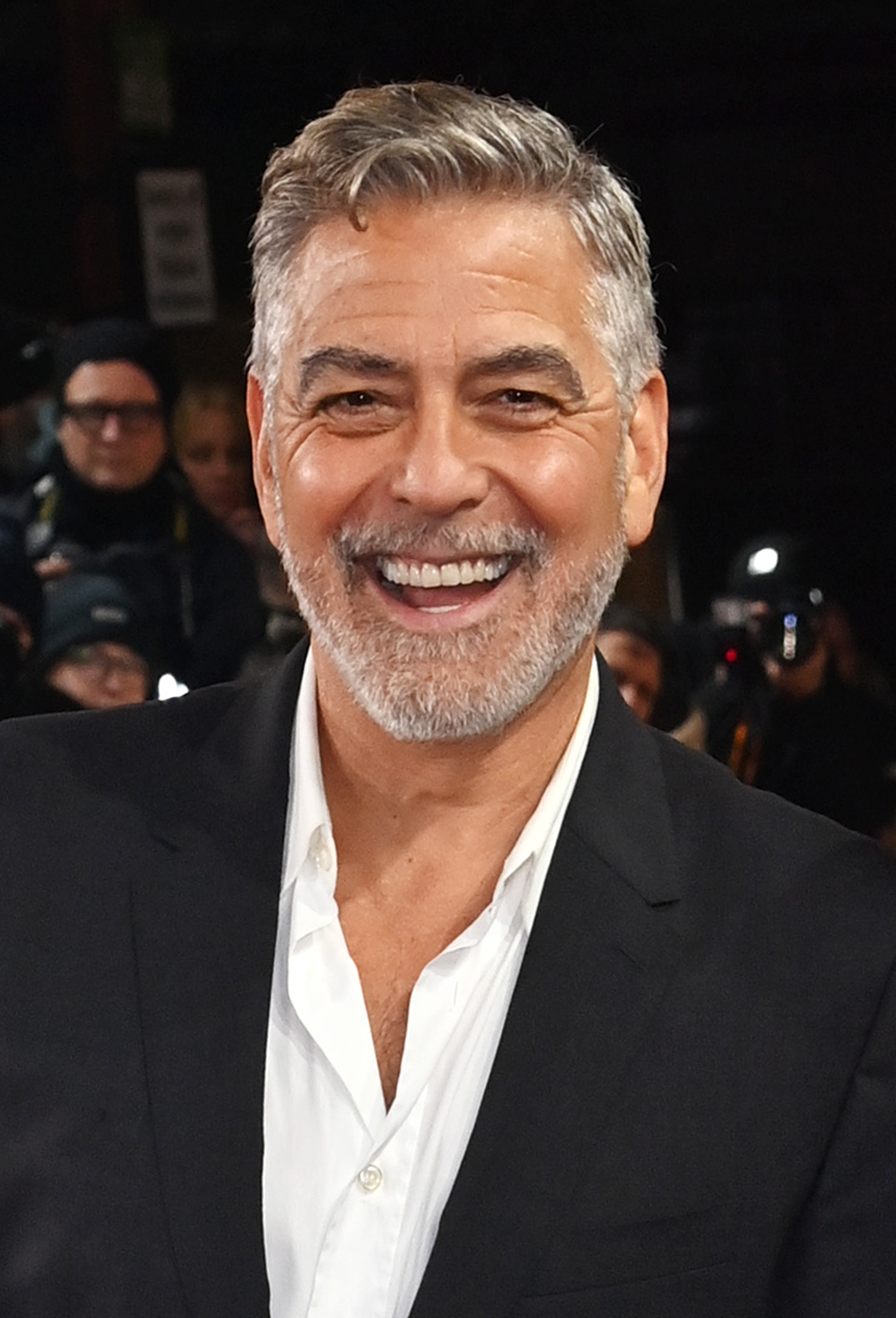 A man with short gray hair and a beard, dressed in a black suit jacket and white shirt, smiles widely at the camera at a public event