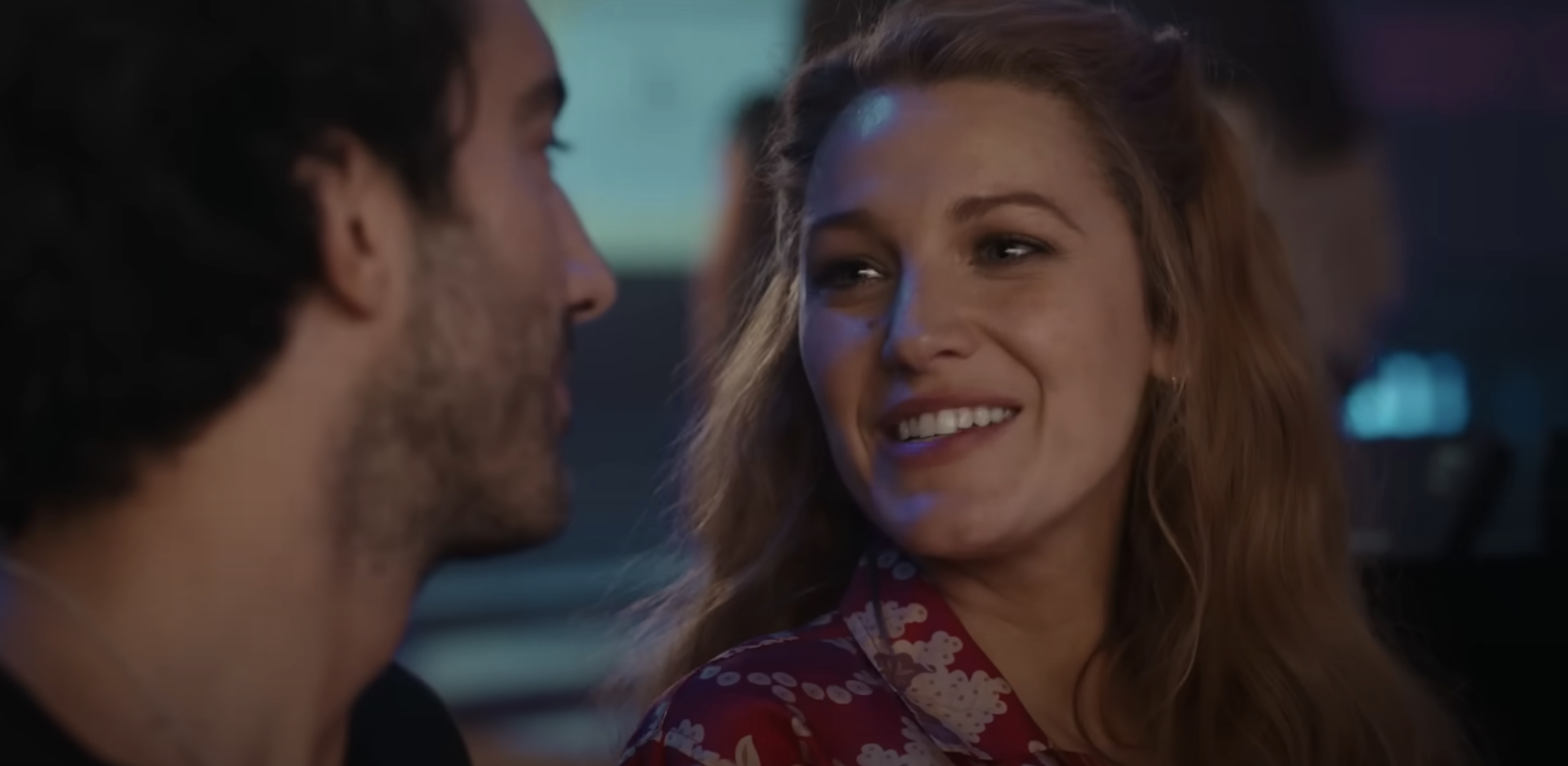 Blake Lively, wearing a floral-patterned top, smiles at a man. The scene appears to be from a movie or TV show