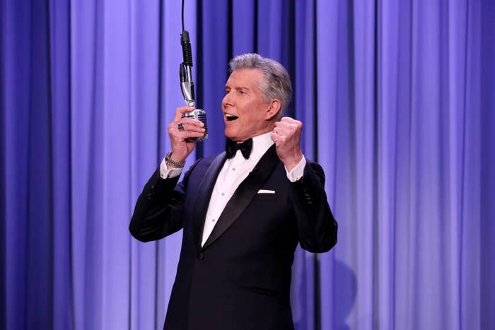 Man in a black tuxedo and bow tie, holding a microphone, poses with one hand closed into a fist in front of a stage curtain. Name unknown