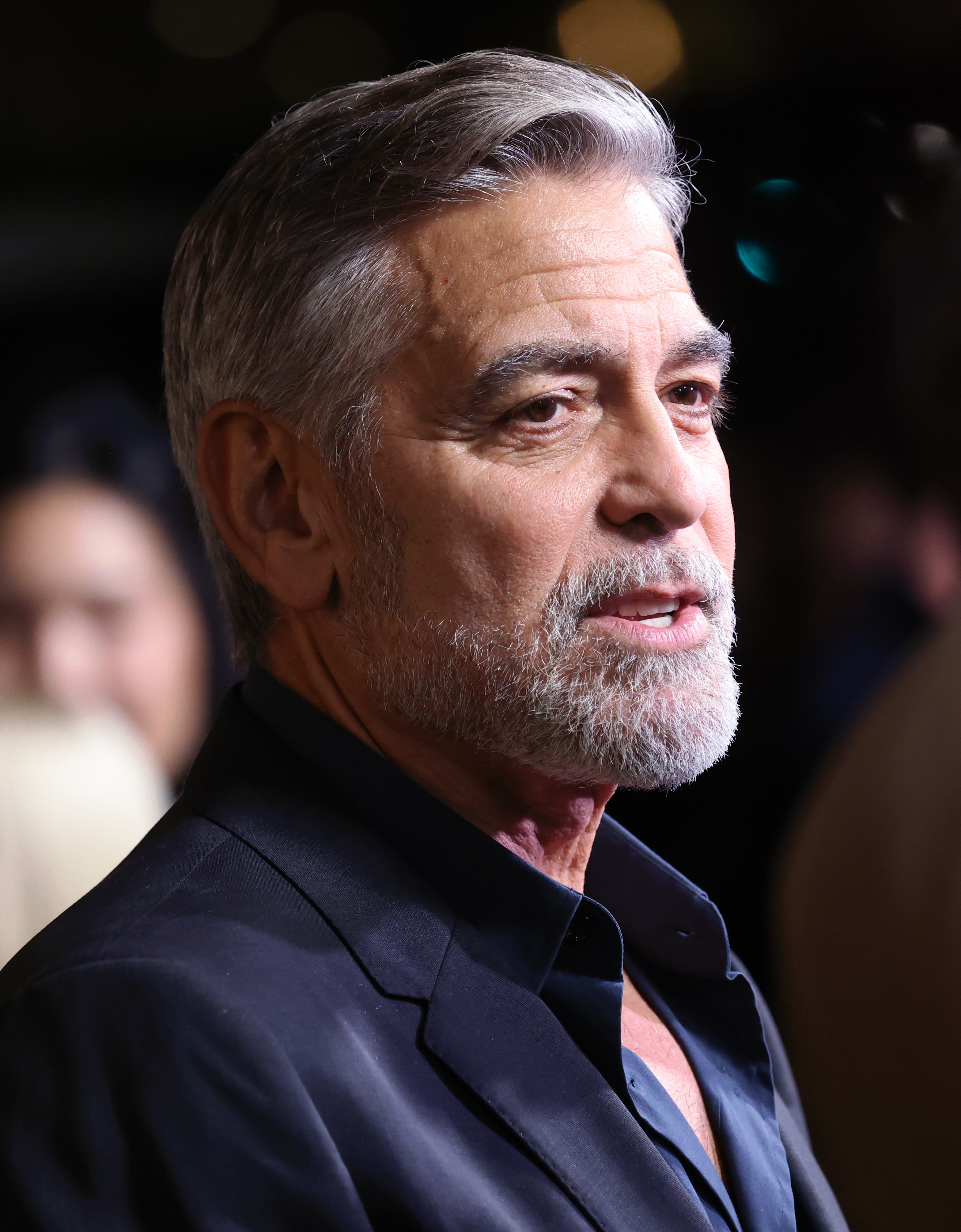 George Clooney, with grey hair and beard, attends an event wearing a dark suit and shirt