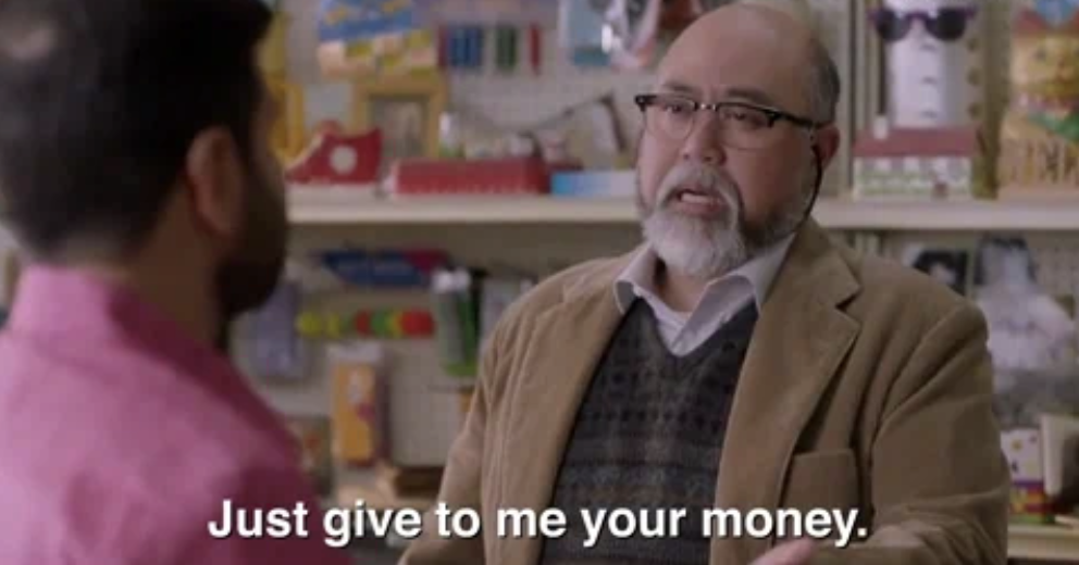 An older man with glasses, beard, and wearing a brown blazer with a sweater instructs &quot;Just give to me your money&quot; to a person whose back is facing the camera