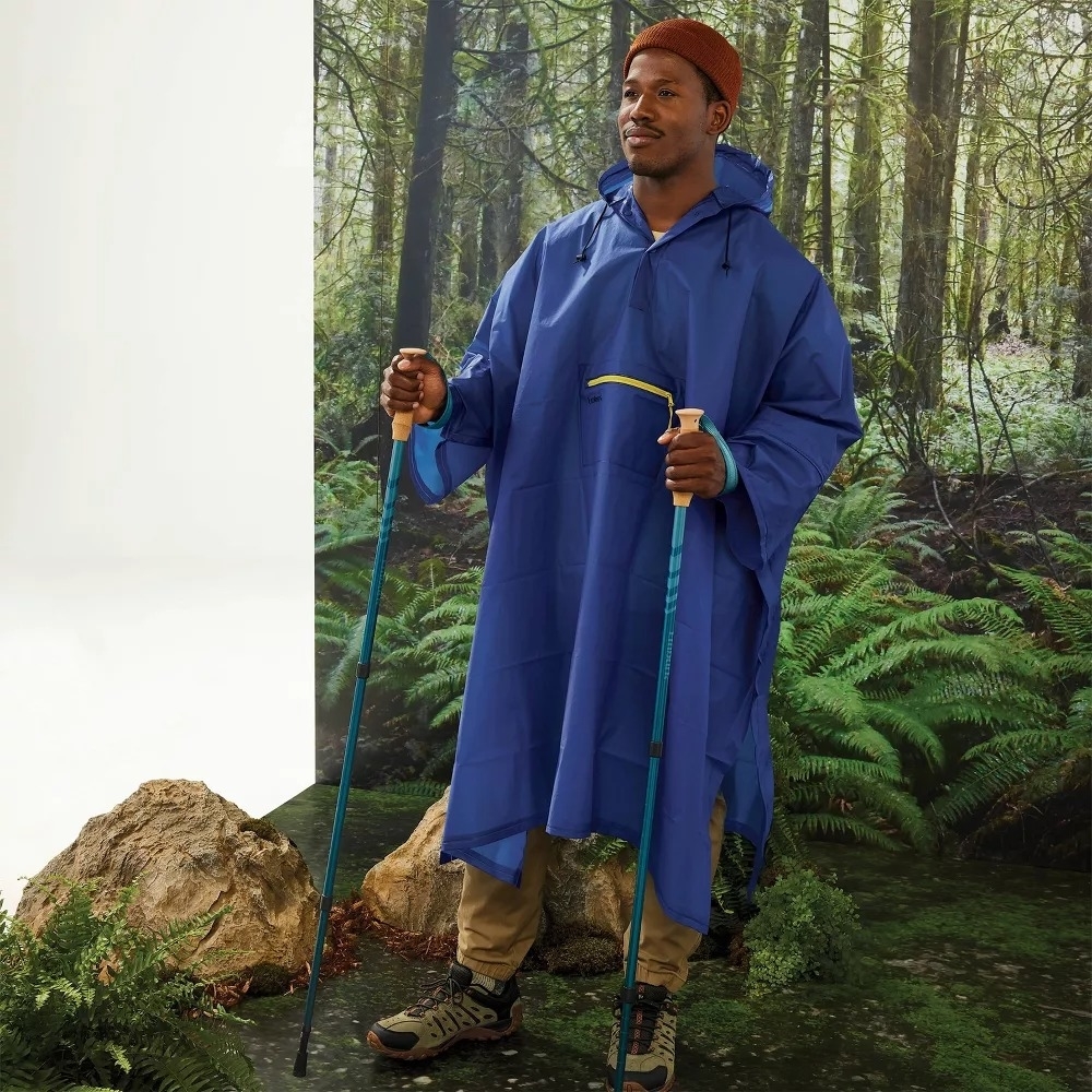 A model in hiking shoes and a knit hat wears the poncho and grips trekking poles.