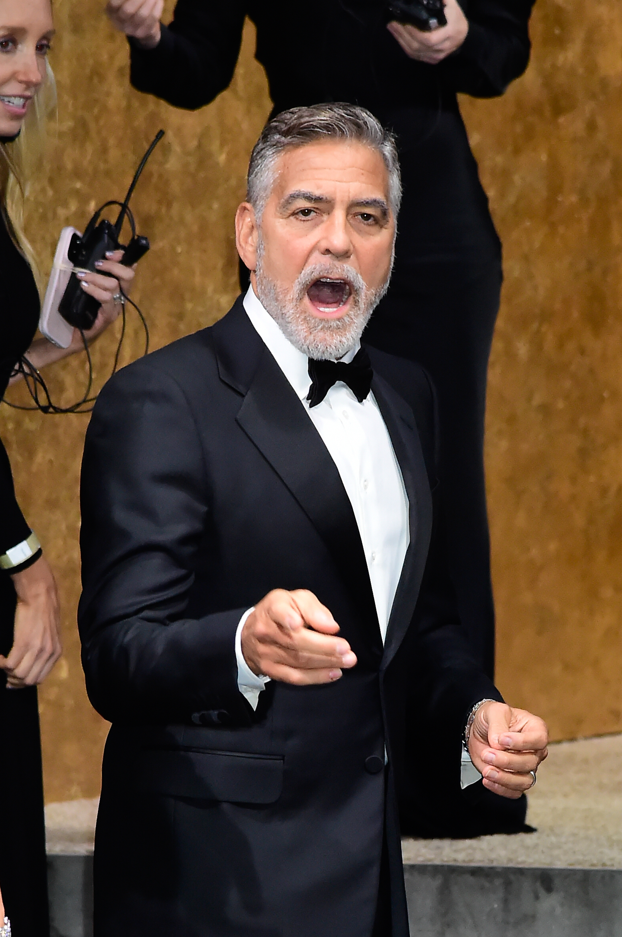 George Clooney, in a classic black tuxedo with a bow tie, gestures while standing at an event. Others around him are in formal attire