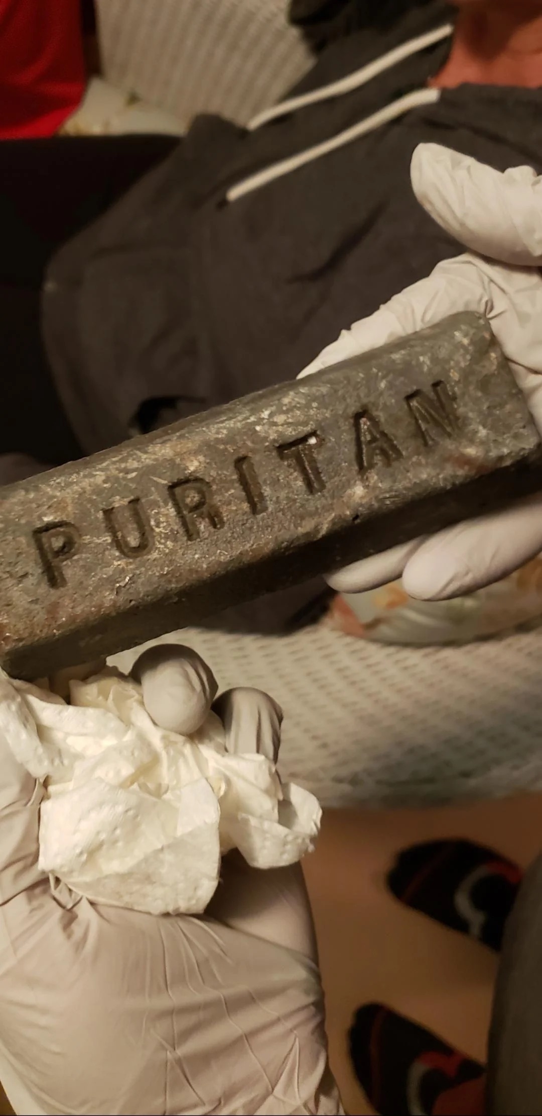 Gloved hands holding a metal bar engraved with the word "Puritan."