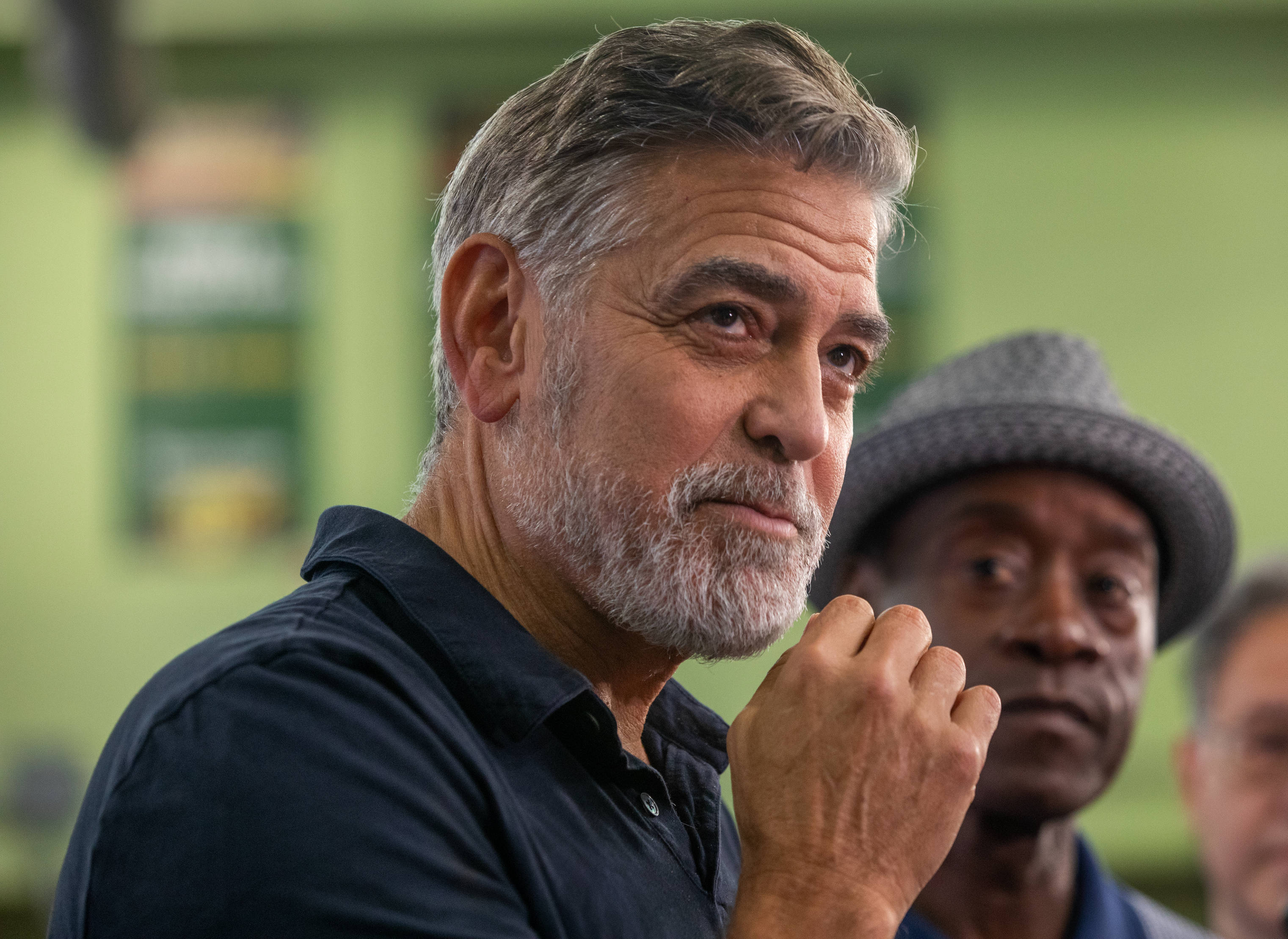 George Clooney and Don Cheadle attend an event; Clooney wears a dark shirt and has a beard, while Cheadle is partially visible behind him wearing a patterned hat