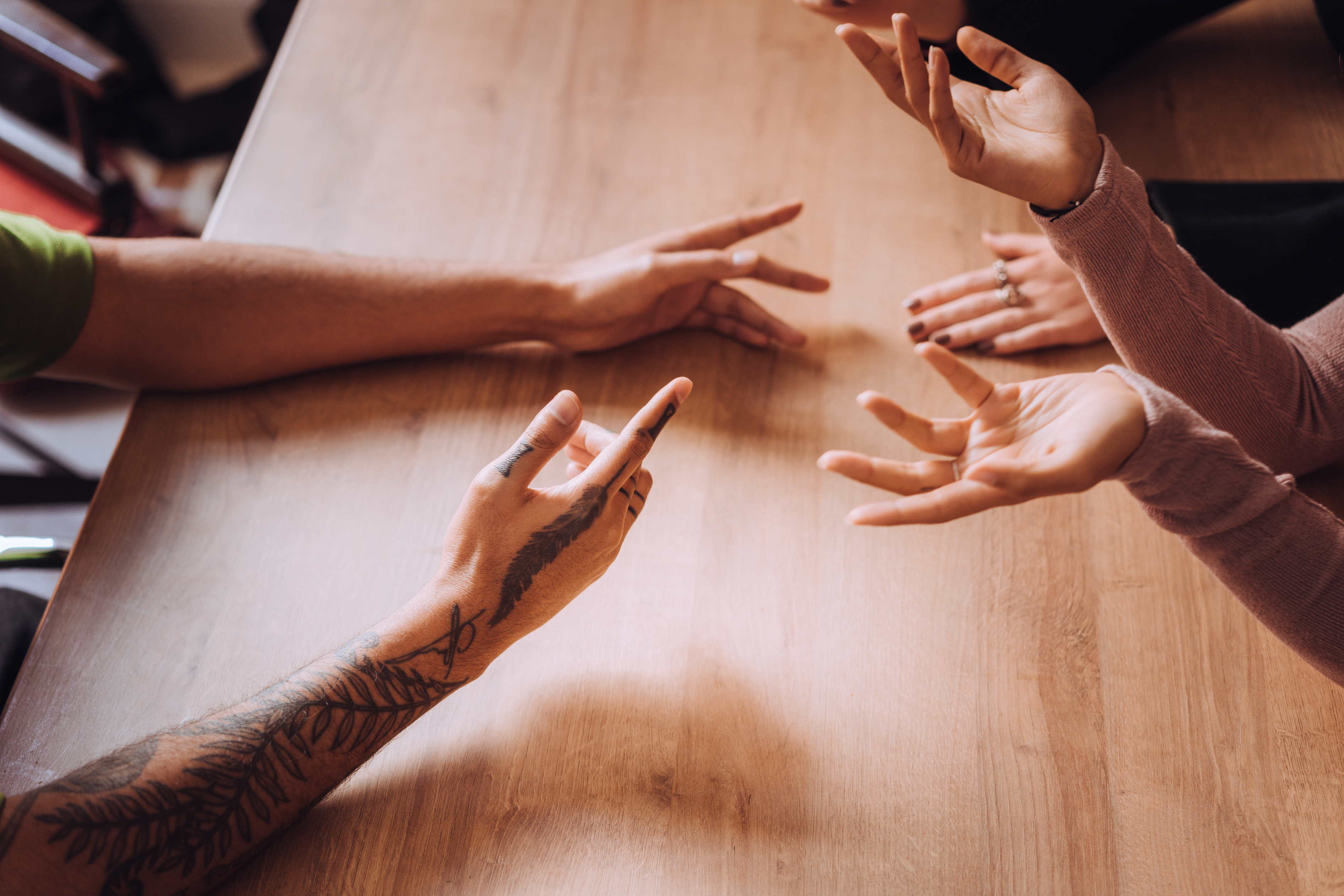 Several people are at a wooden table, using hand gestures during a discussion. One person has a tattoo on their arm