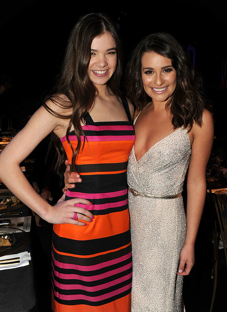 Hailee Steinfeld and Lea Michele pose together. Hailee wears a striped dress, and Lea wears a deep V-neck dress cinched at the waist