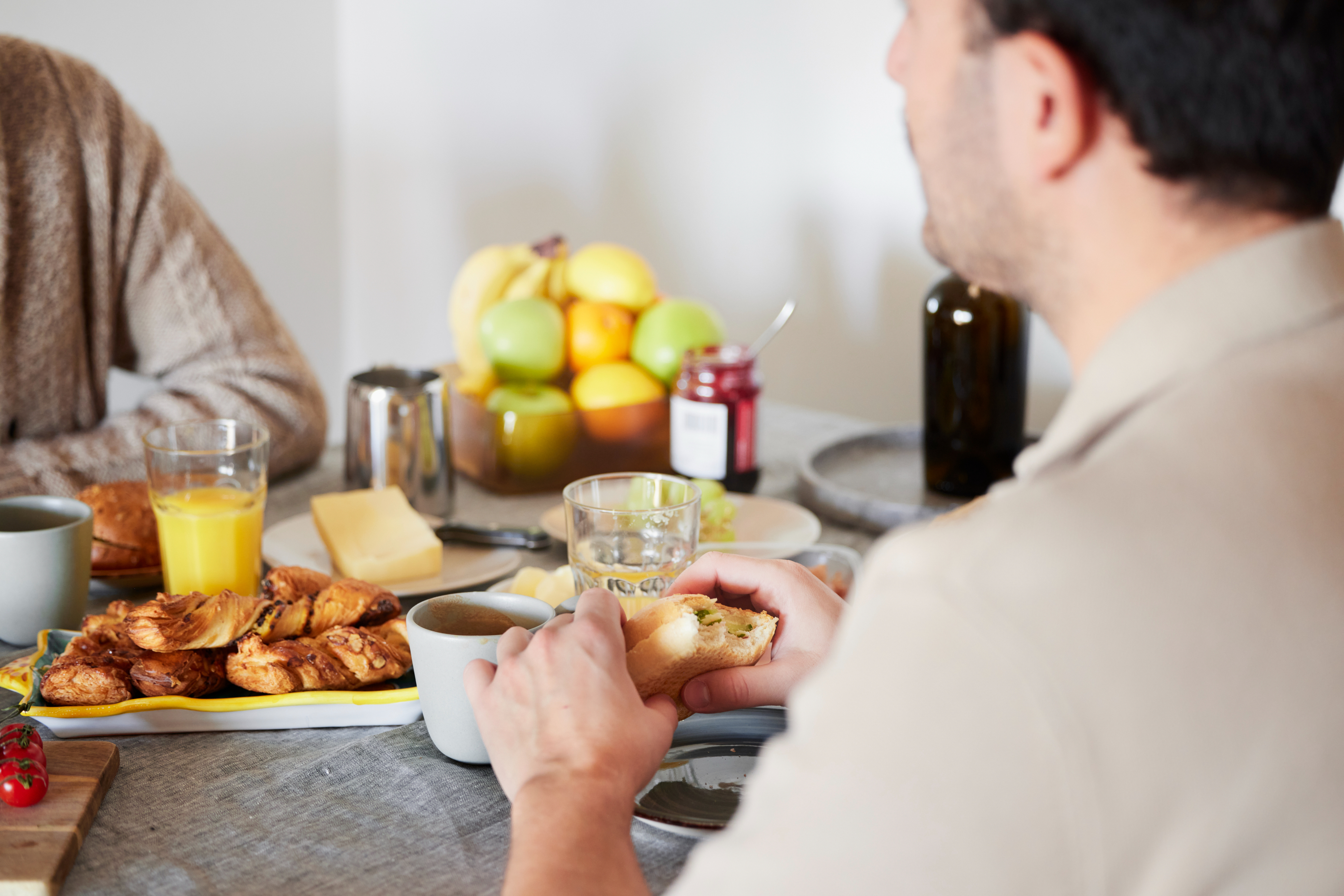 Two people sharing a breakfast table with croissants, fruits, orange juice, coffee, and various jams. Faces are not visible