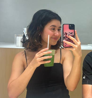 A woman takes a mirror selfie holding a green smoothie and smiling, wearing a black tank top