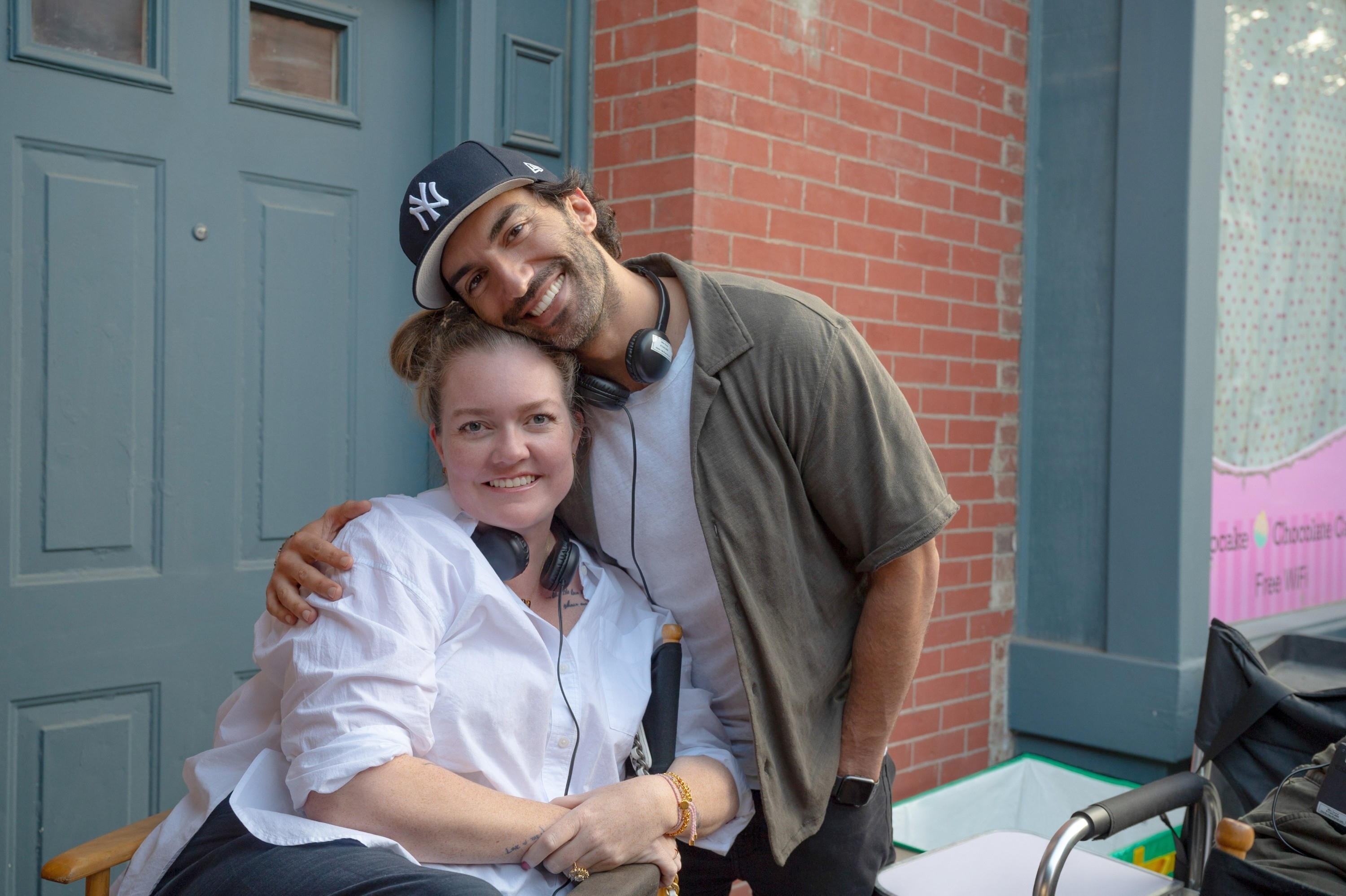 Two people smile and pose together; one sitting in a wheelchair and the other standing with an arm around their shoulder. Both wear headsets and casual clothing