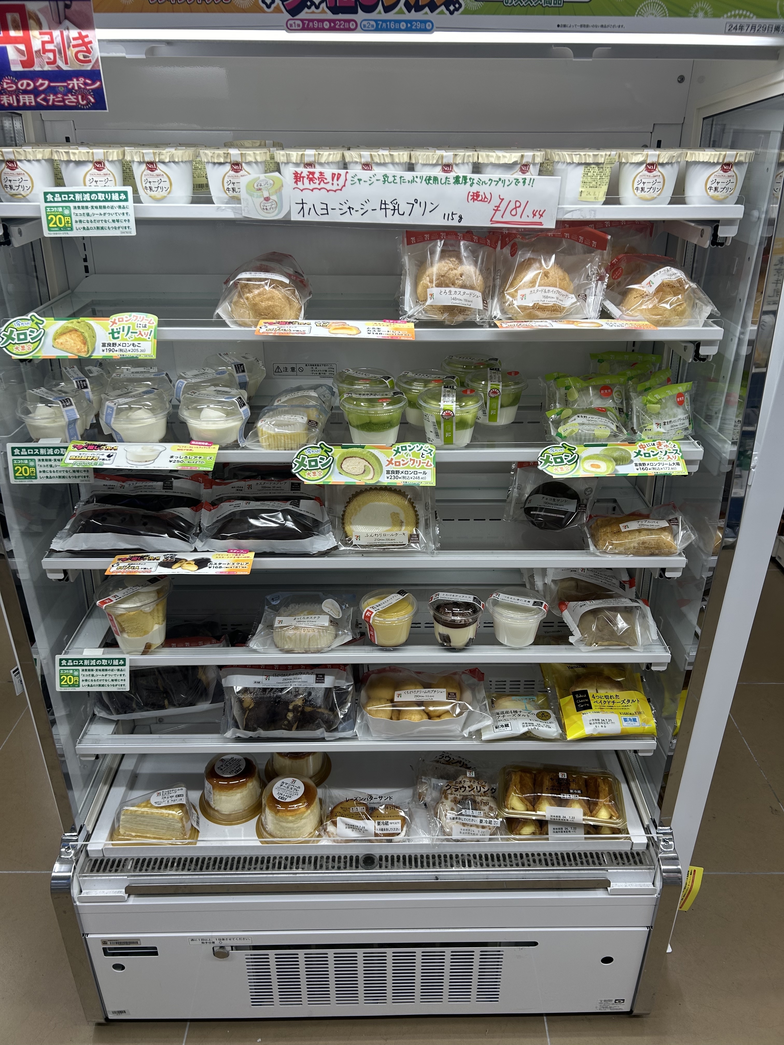 A refrigerated display case filled with various packaged desserts, including puddings, cakes, and pastries. All items have labels primarily in Japanese