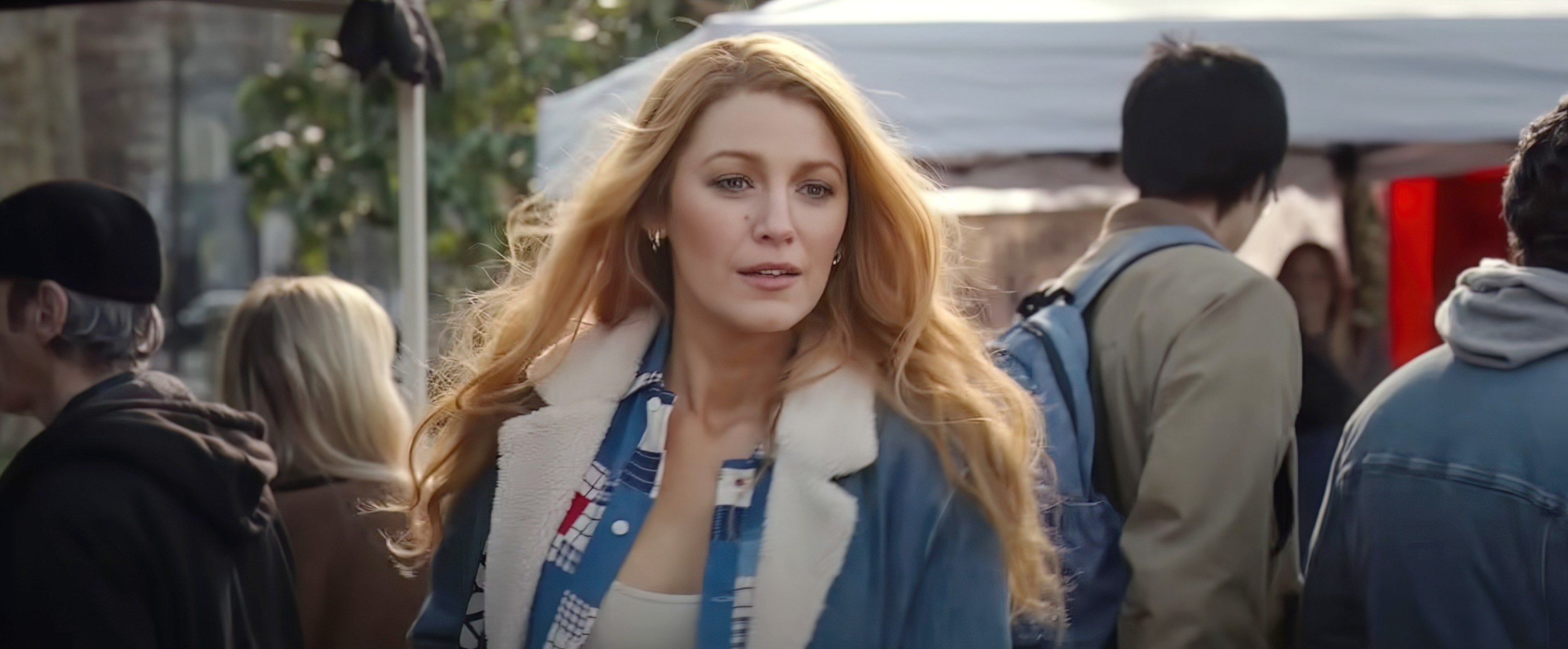 Blake Lively is in an outdoor market scene, wearing a plaid shirt, a denim jacket with a shearling collar, and walking among other people