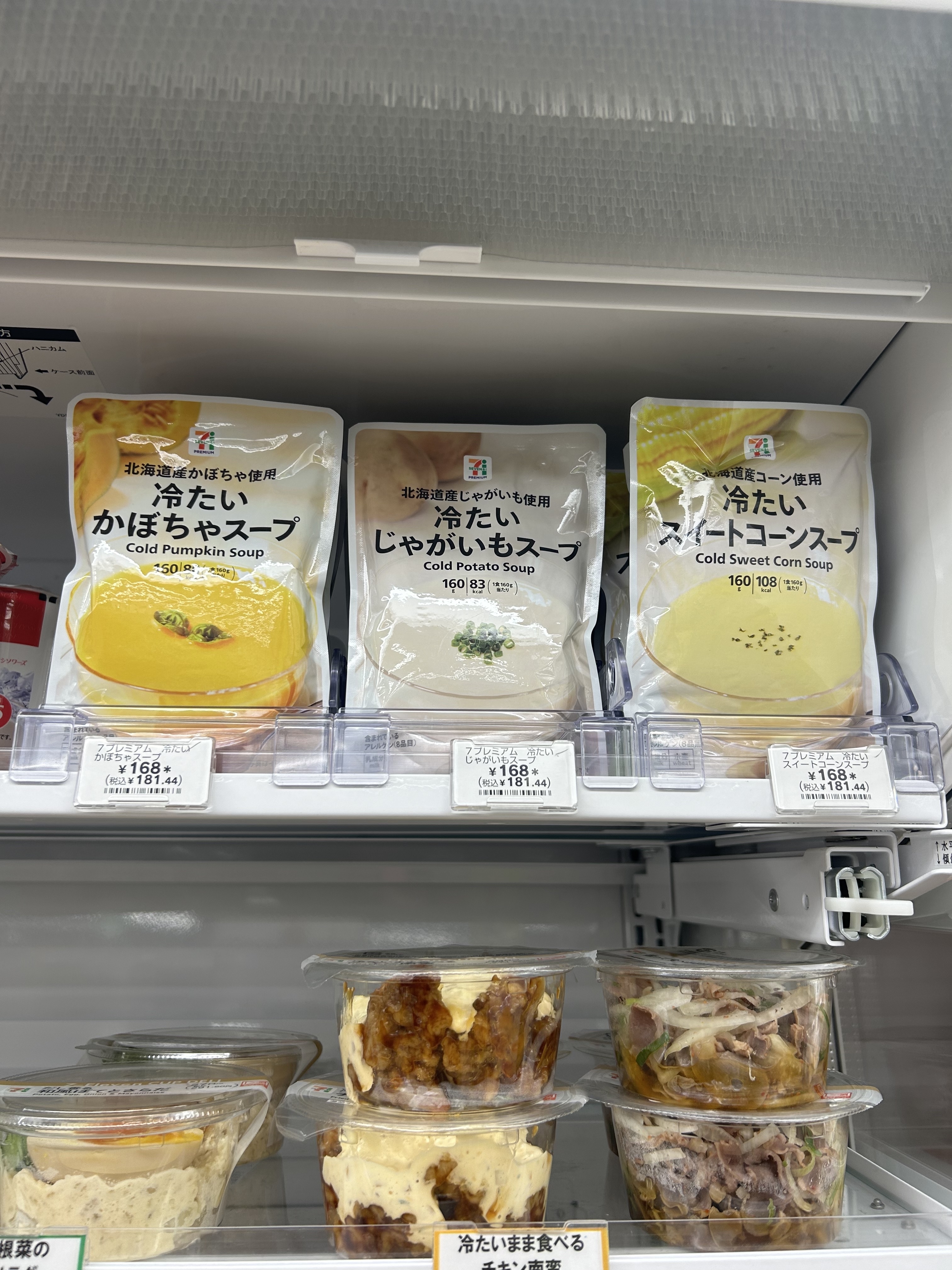 Three packaged cold soups (cold pumpkin, potato, and sweet corn) on a store shelf, with various pre-packaged food items displayed below them